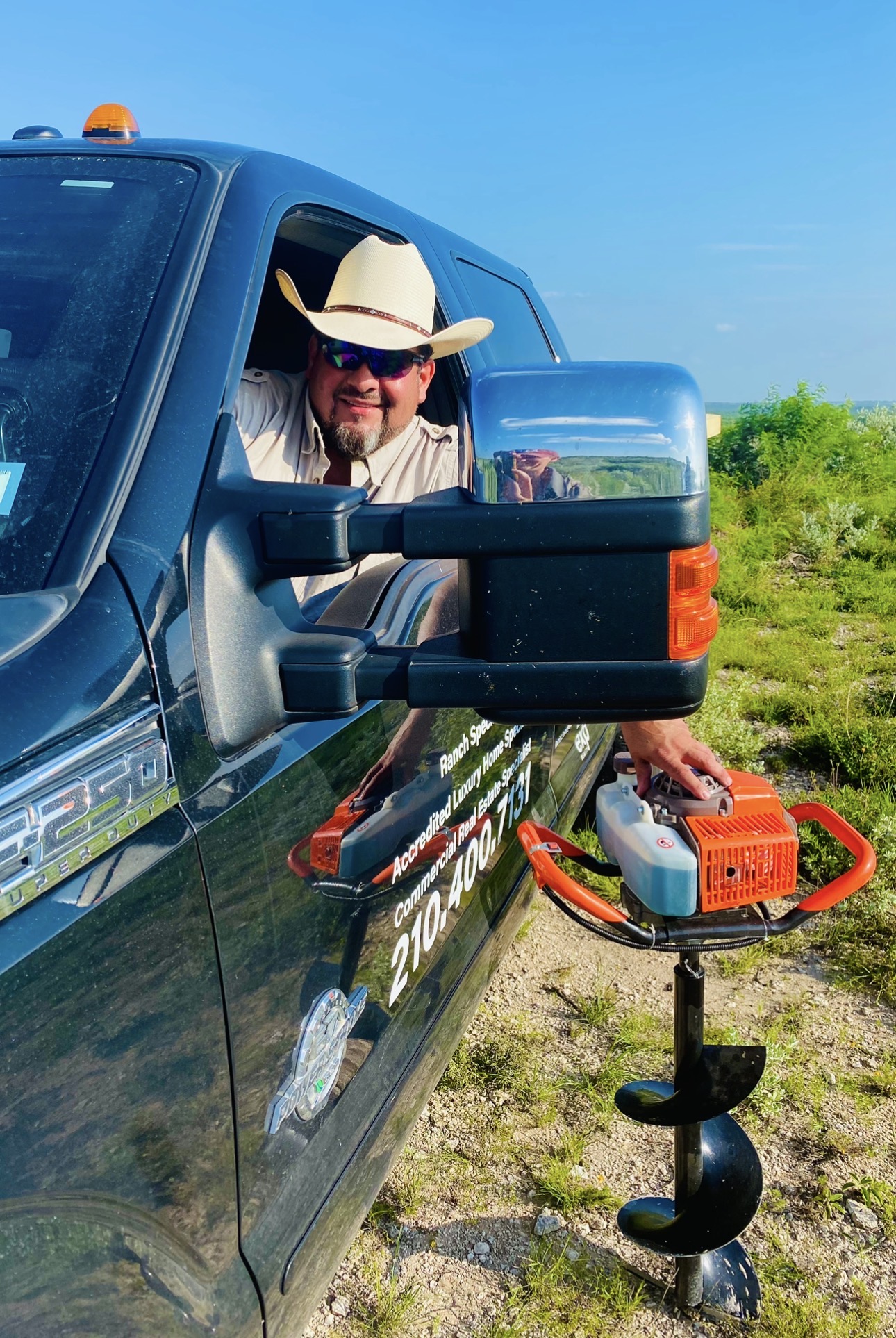 Man wearing a cowboy hat and sunglasses leaning out of a blue truck window, with an orange and black auger on the ground nearby.
