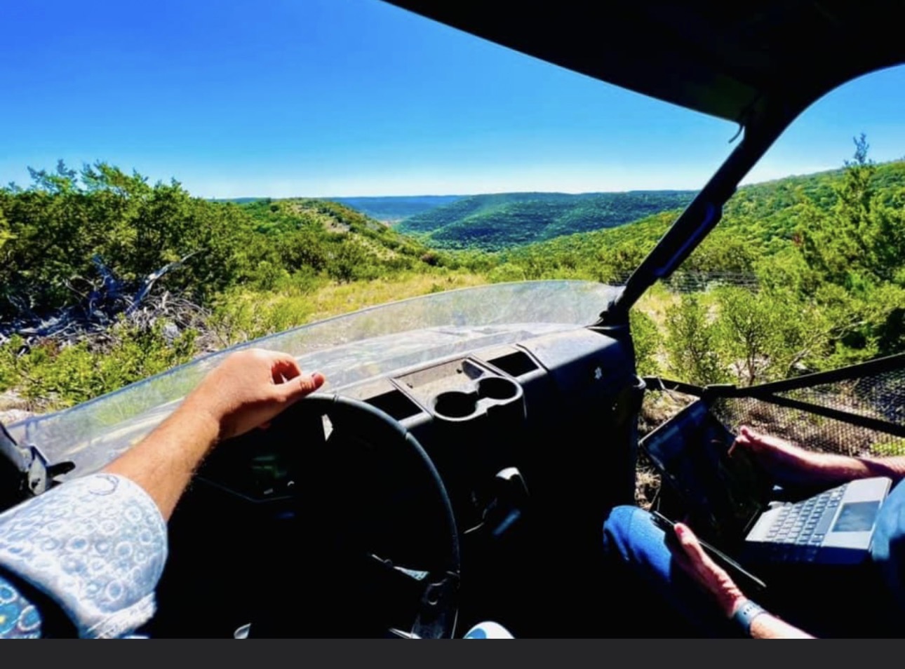 View from inside a vehicle showing a dashboard, steering wheel, and two people, with a landscape of green hills and blue sky outside