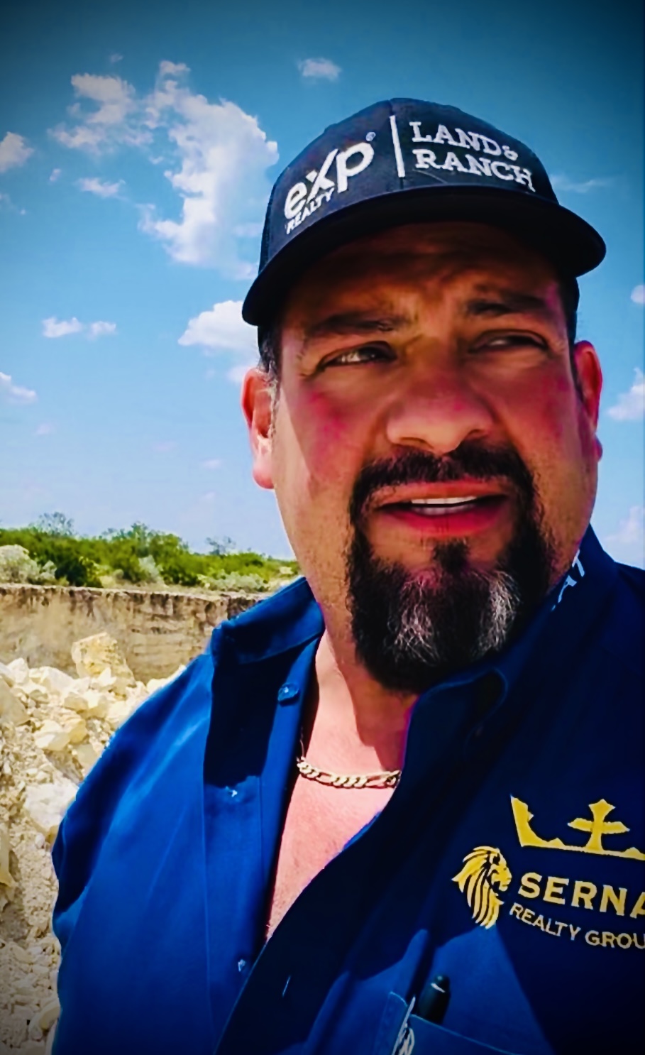 Man outdoors wearing a cap and blue shirt, with a partly cloudy sky in the background.