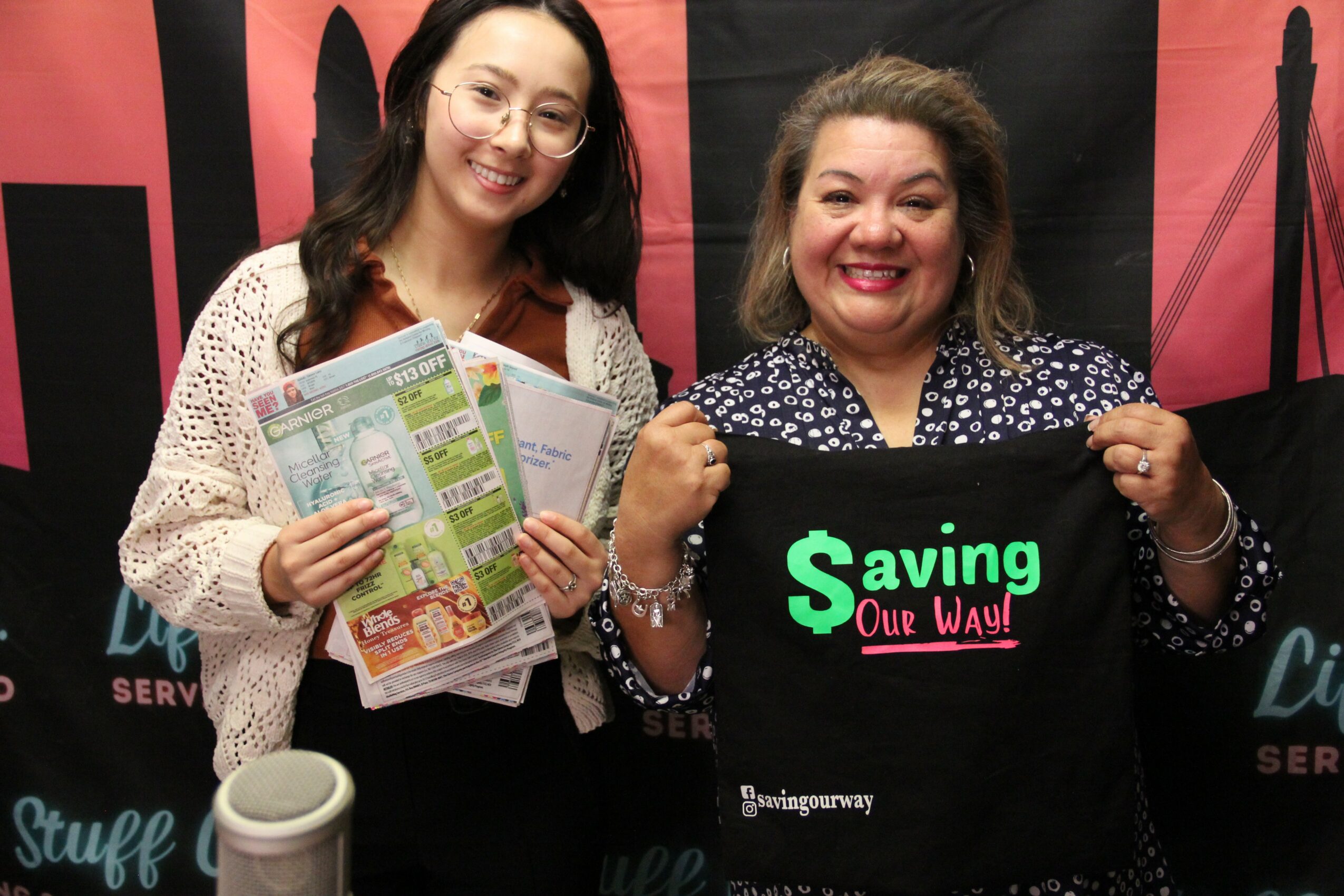 Two women smiling, holding flyers and a black bag with green and pink text, standing in front of a black and pink backdrop.