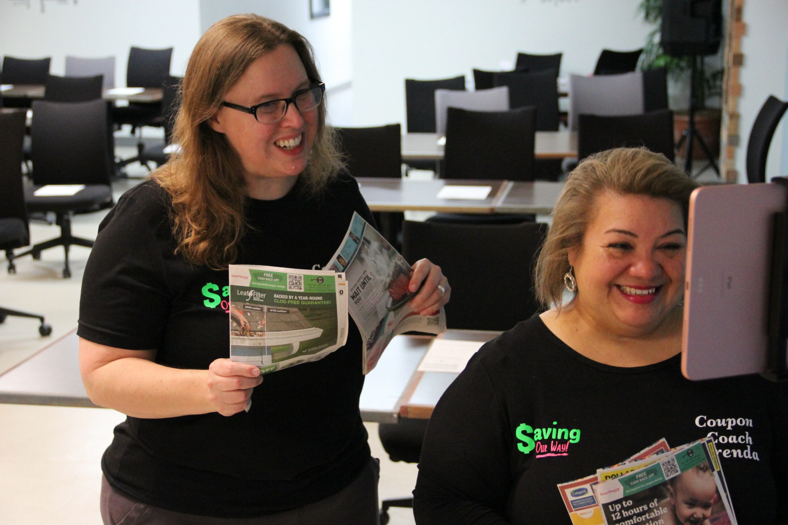 Two women smiling and looking at a computer screen in a conference room, holding pamphlets, with chairs and tables in the background.