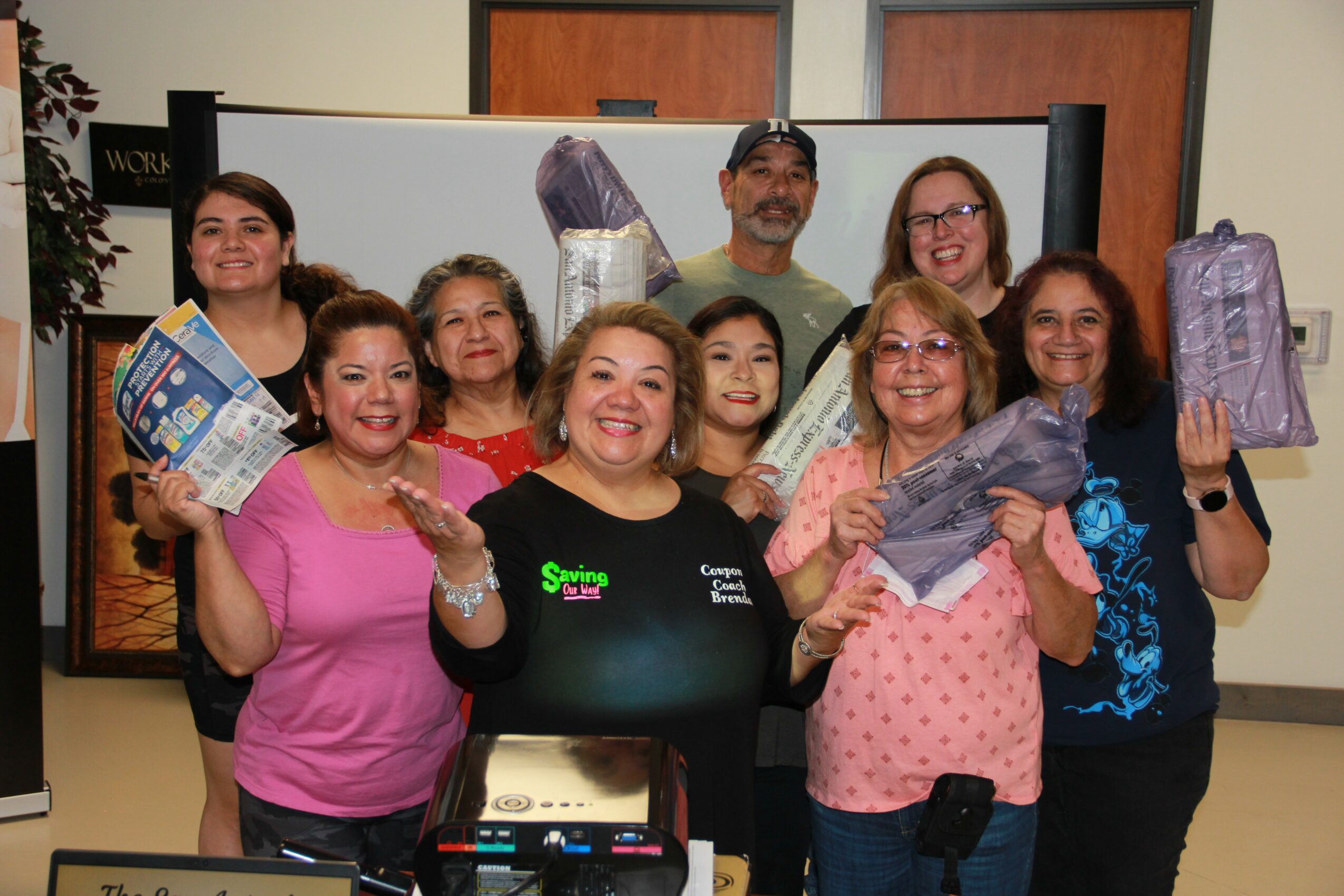 Group of nine people smiling and holding wrapped gifts in a room with a large screen behind them.
