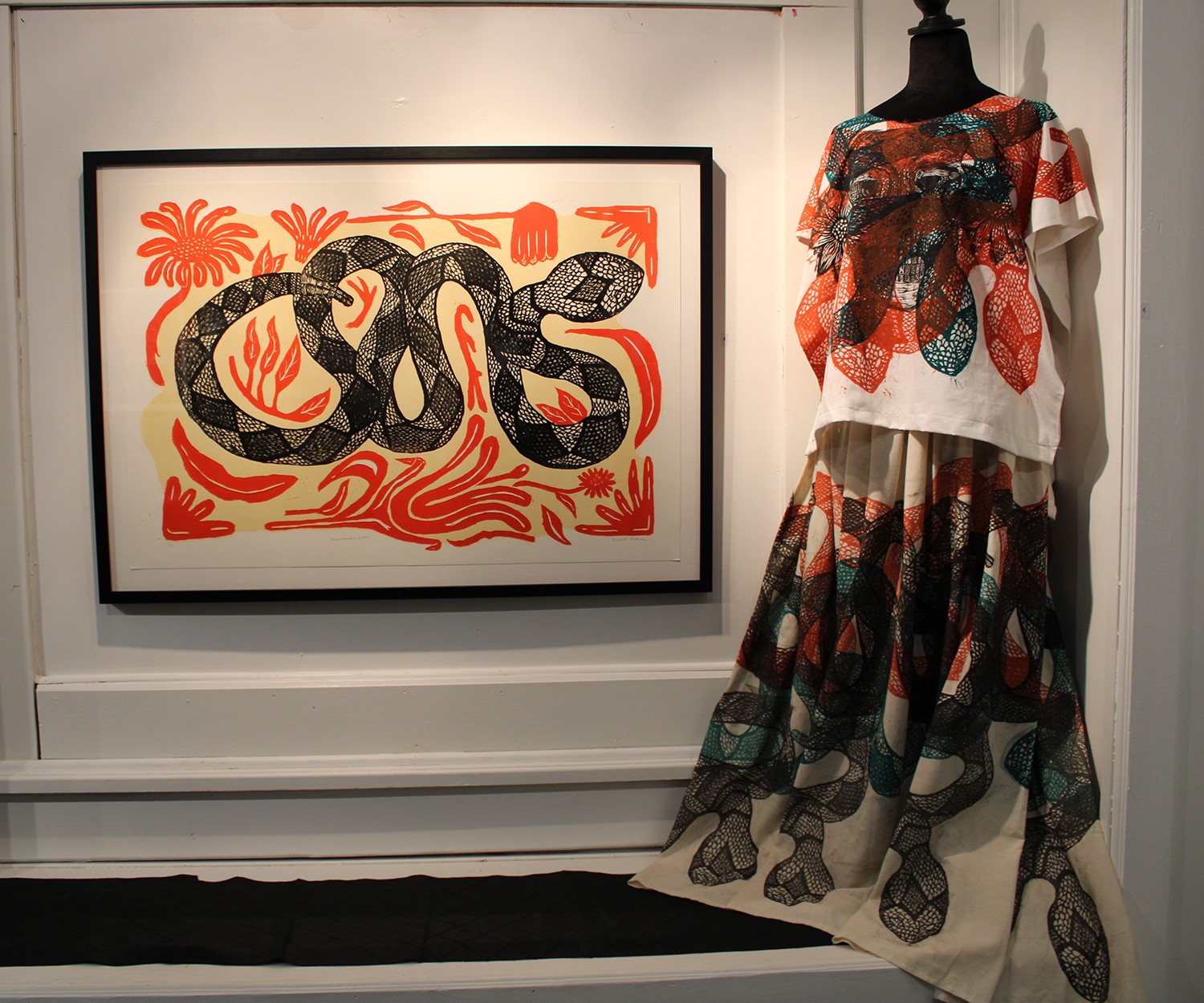 A framed artwork of a snake with red and black patterns hangs on a wall, next to a mannequin dressed in a colorful, patterned outfit.