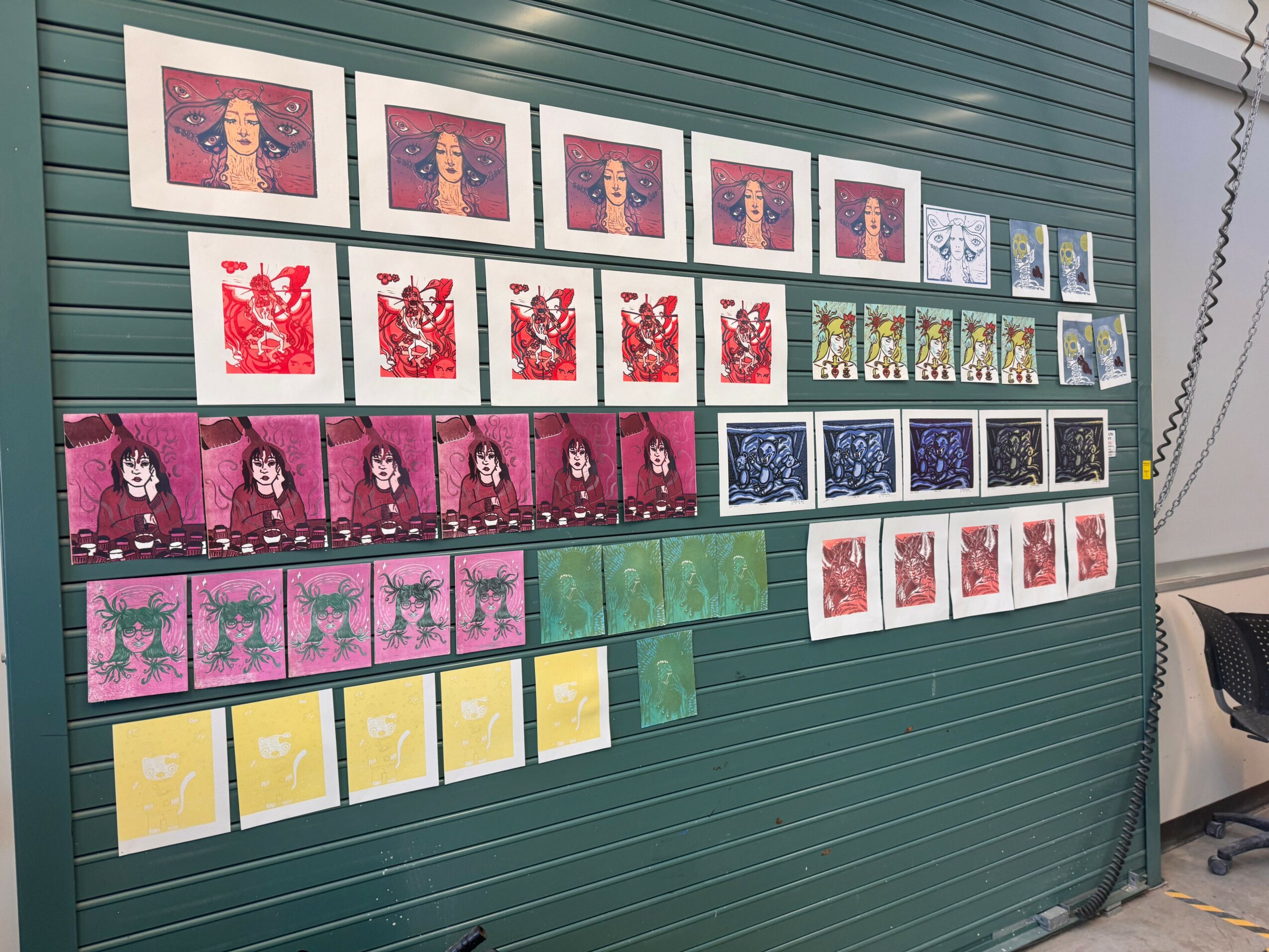 Display of colorful artwork and prints arranged on a green wall, including portraits, abstract designs, and patterned images.