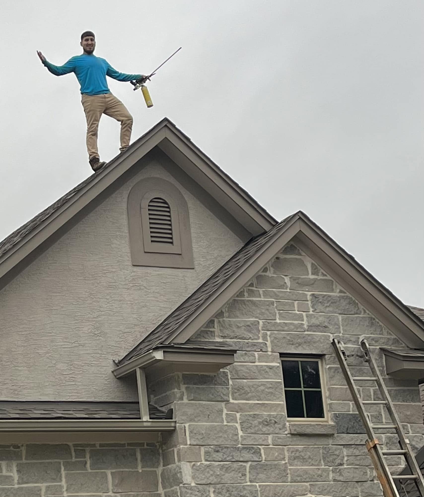 Person standing on roof peak holding a spray bottle, overcast sky background.