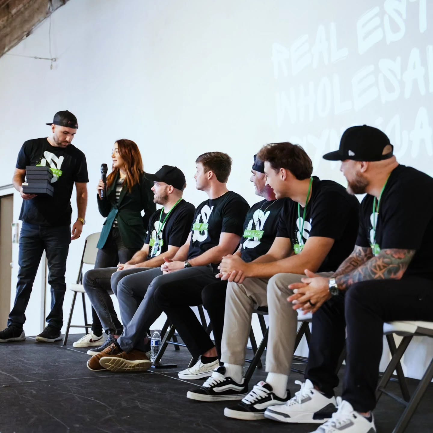 Six people sitting on chairs on stage, one person standing and speaking, a large screen in the background, casual clothing, some with tattoos.