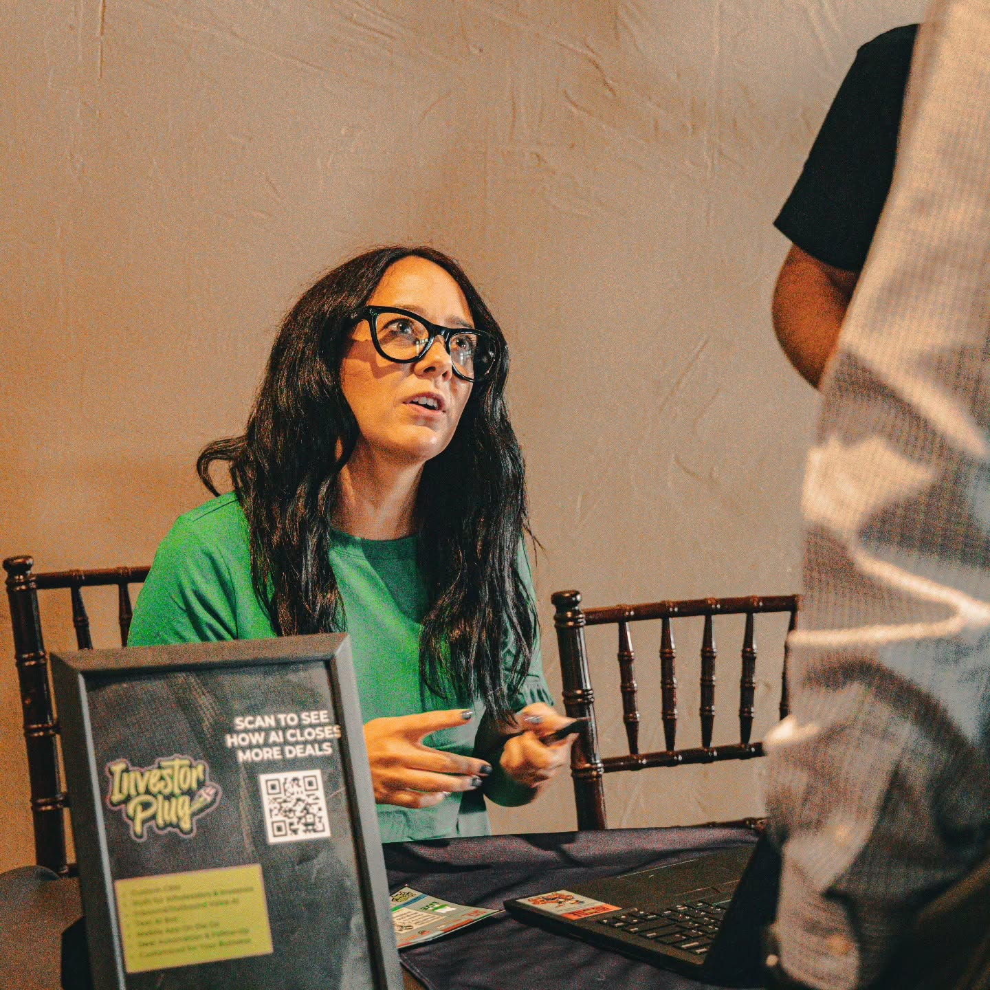 Woman with long dark hair and glasses wearing a green shirt, sitting at a table with a laptop and a sign, looking at a person standing nearby.