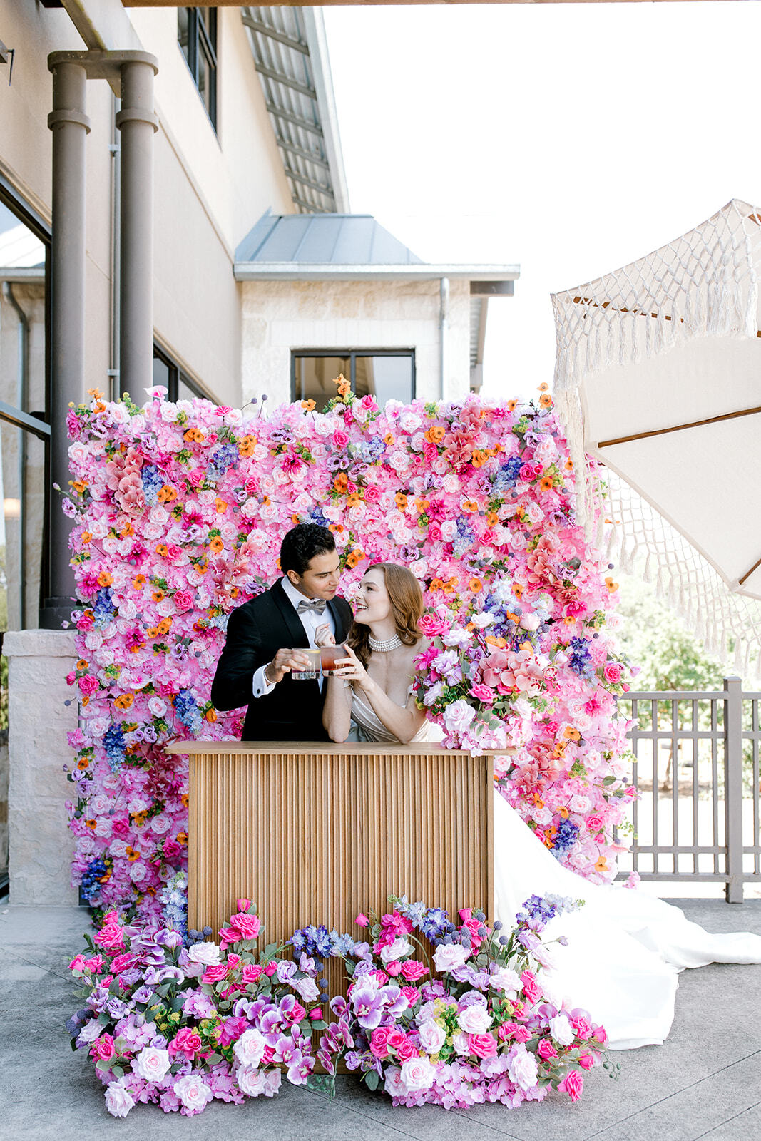 Two people stand behind a wooden table in front of a large pink and purple flower wall, outdoors.