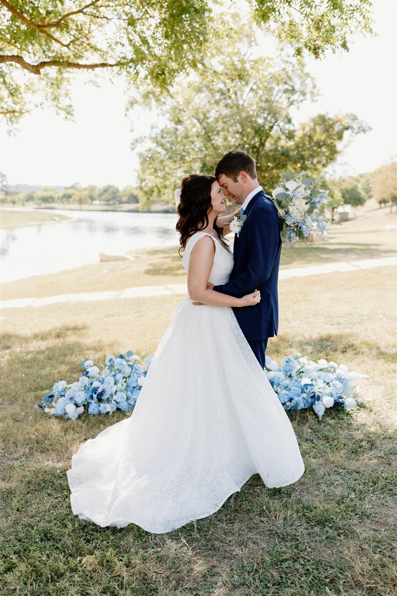 Couple in wedding attire kissing outdoors near a lake with trees and flowers, sunlight filtering through leaves.
