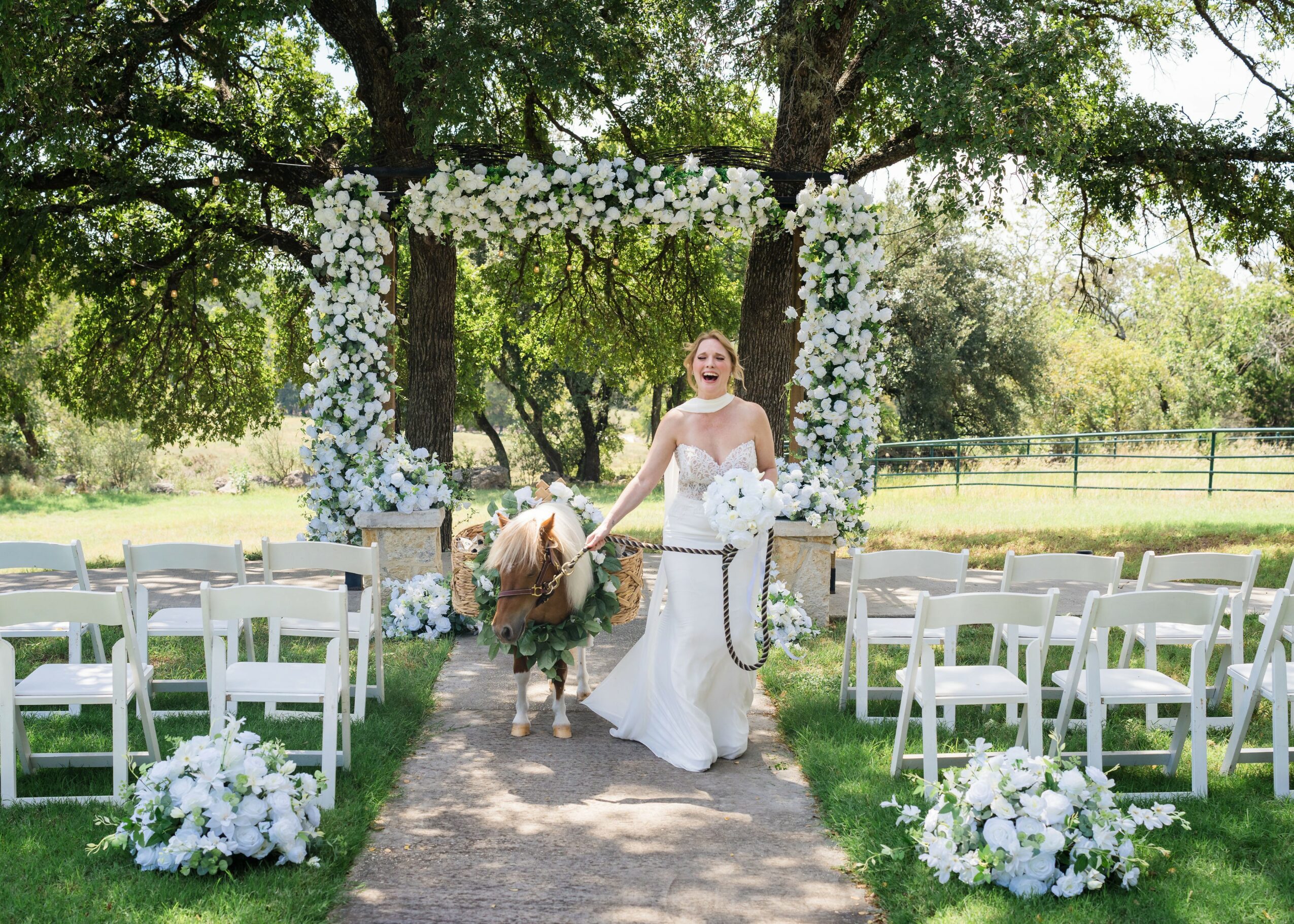 Bride in white dress holding a dog, standing under a floral arch, with chairs and floral arrangements outside.