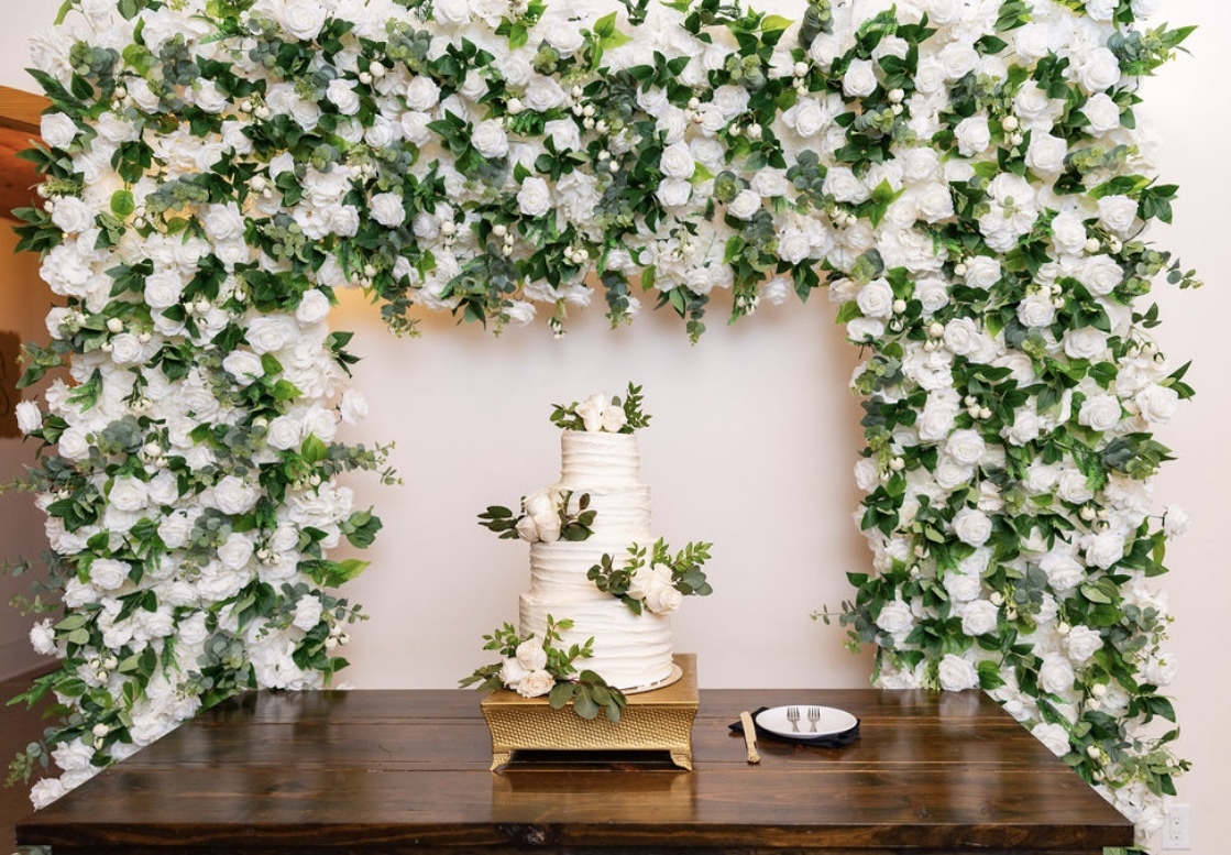 Wedding cake with flowers under a floral arch on a wooden table, with a small dish nearby.
