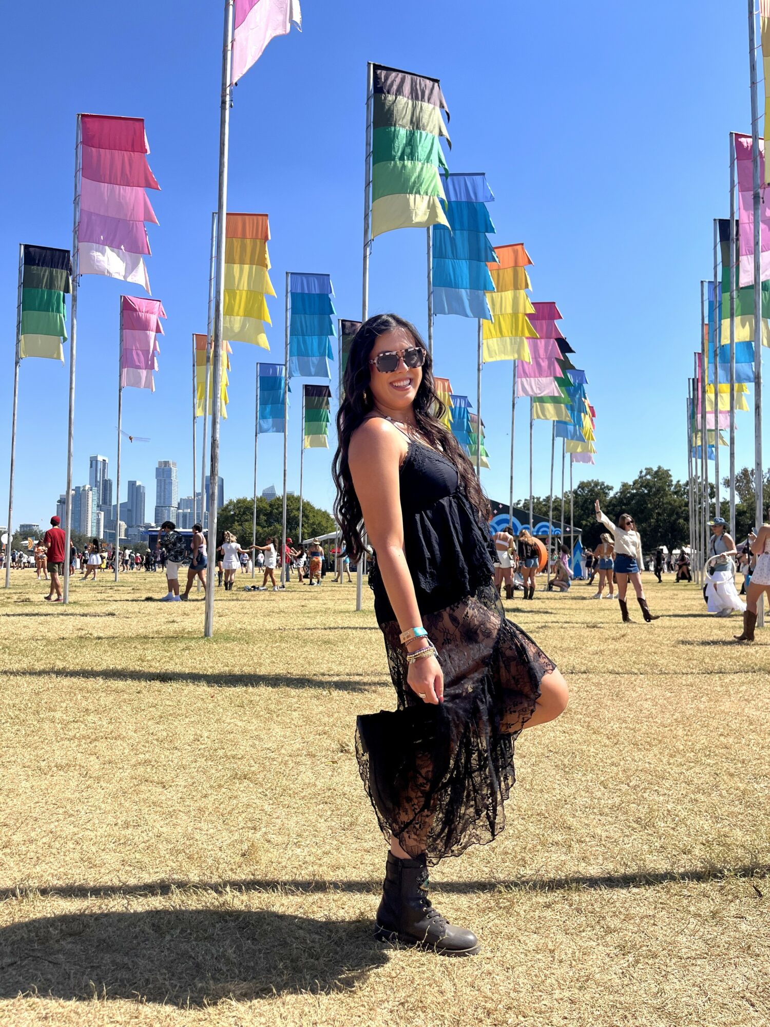 Woman in black dress and sunglasses standing outdoors with colorful flags and city skyline in background.