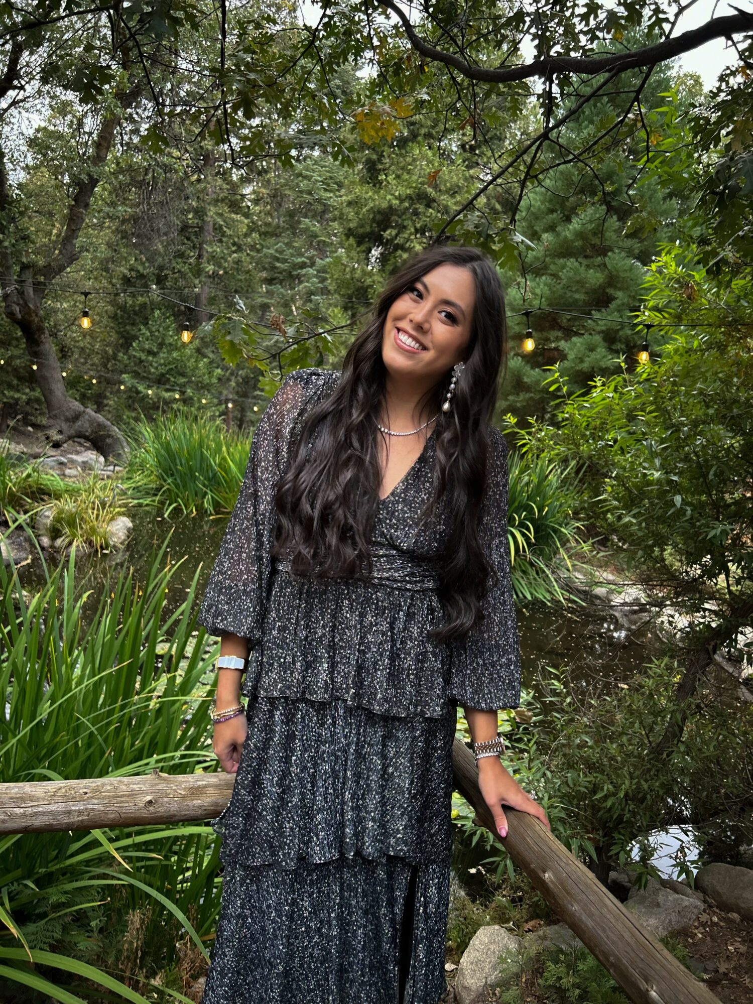 Young woman with long dark hair smiling outdoors in a garden with trees and greenery.