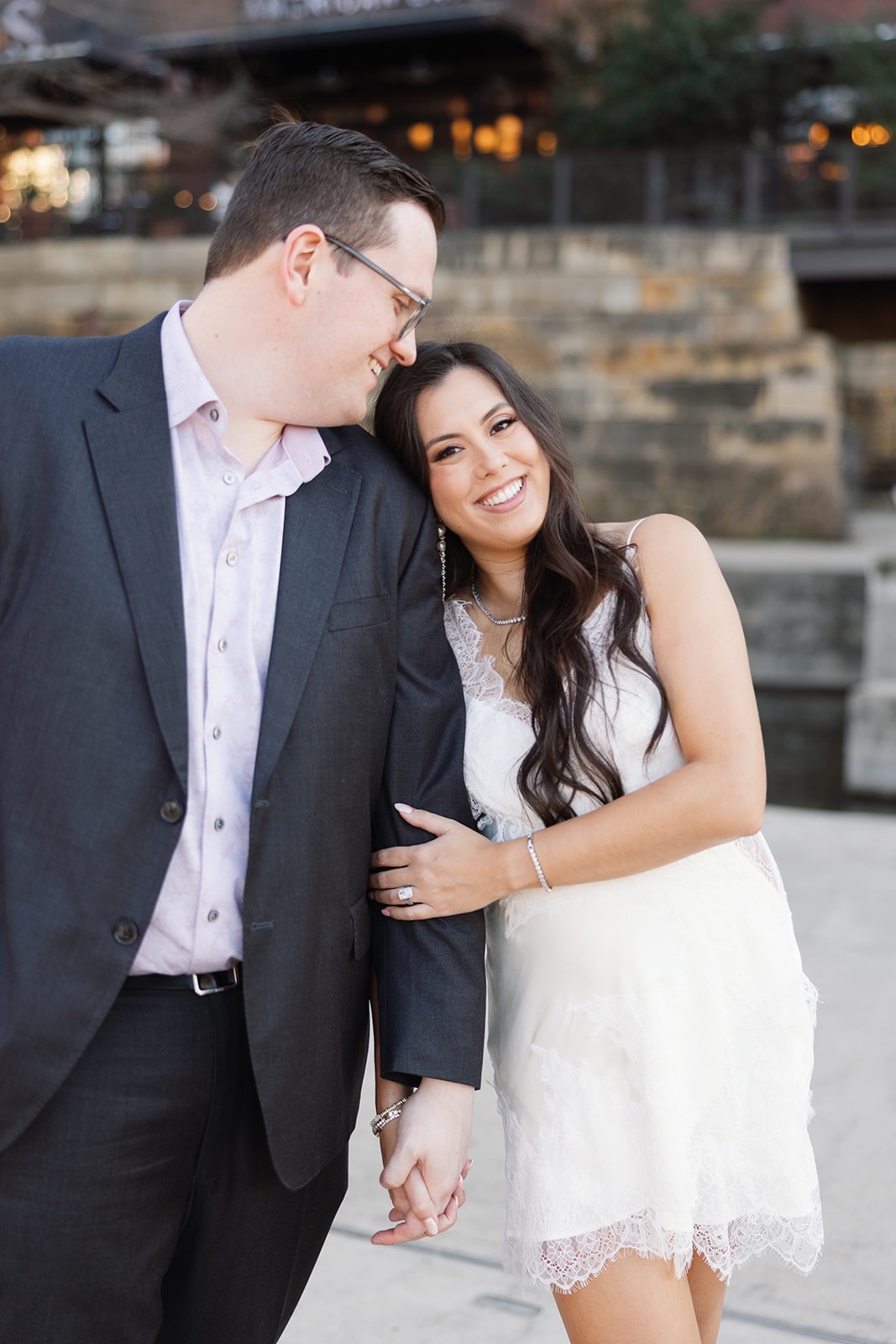 A smiling woman in a white dress leans on a man in a dark suit, holding hands outdoors near a stone wall.