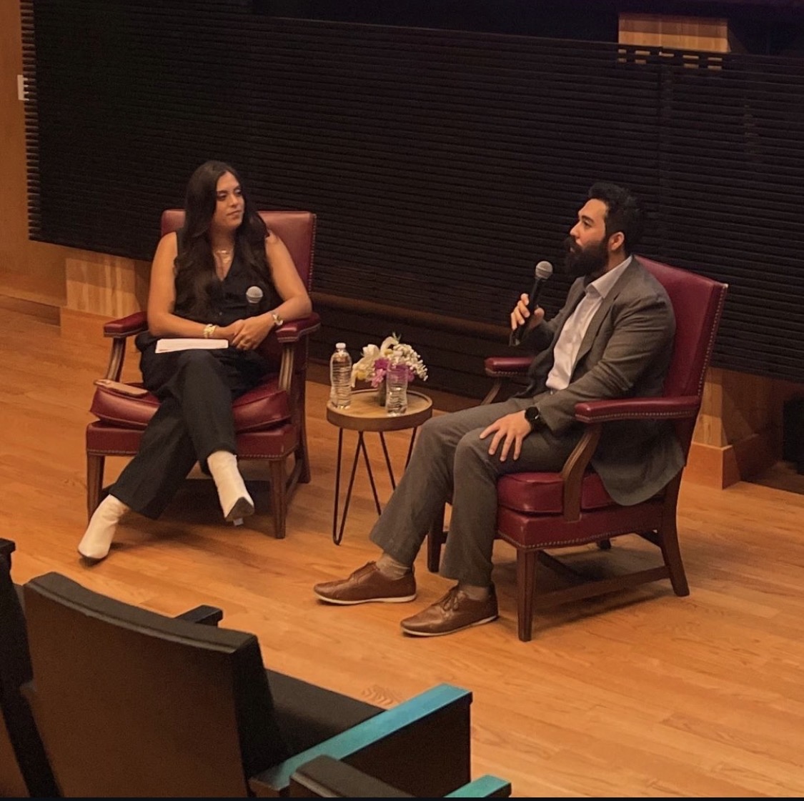 Two people seated in chairs on stage, engaged in conversation, with a small table holding water bottles and flowers between them.