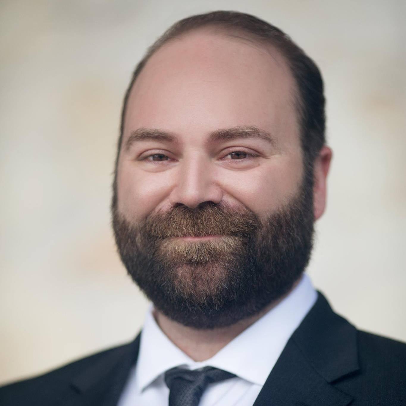 Portrait of a man with a beard, smiling, wearing a suit and tie, against a neutral background.