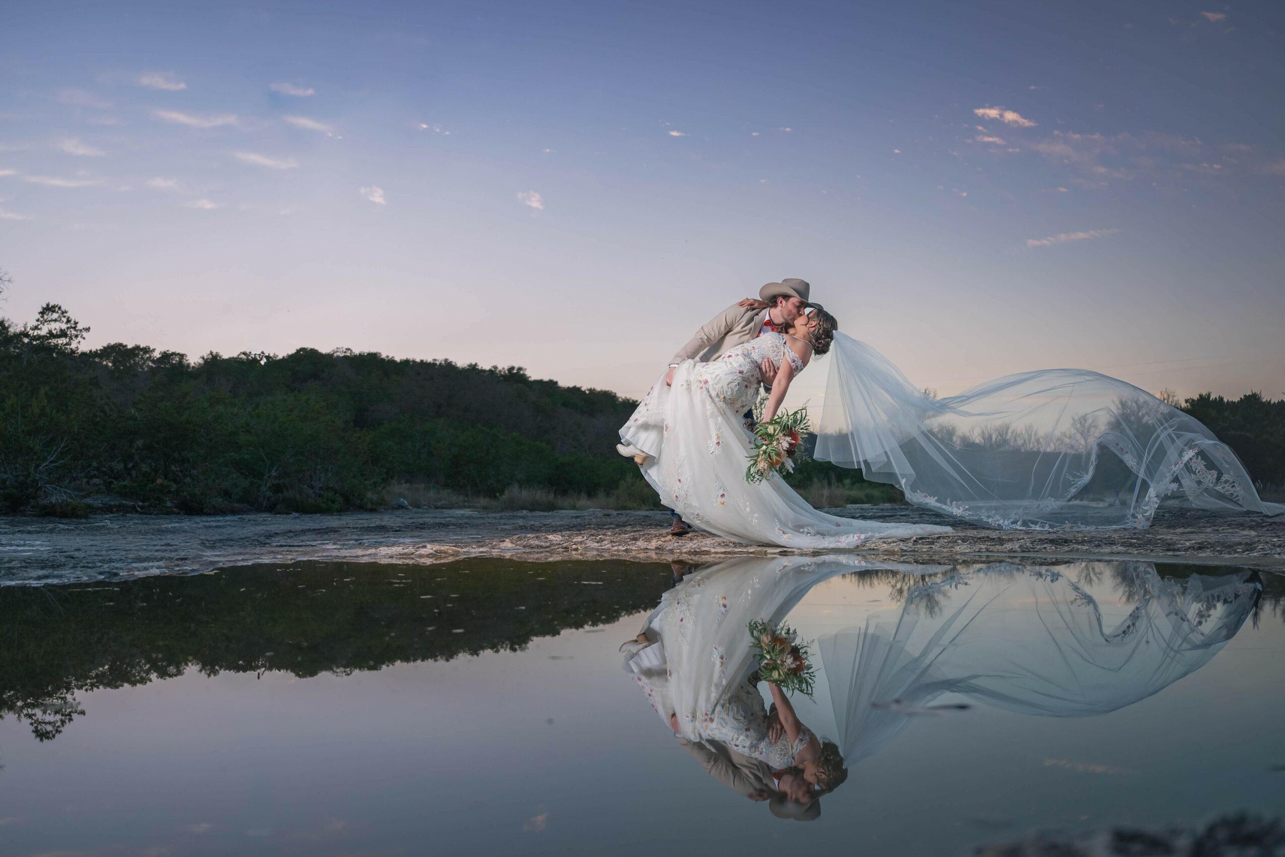 Bride in a white dress and veil kneels by water, holding a bouquet, with her reflection visible in the water, under a twilight sky.