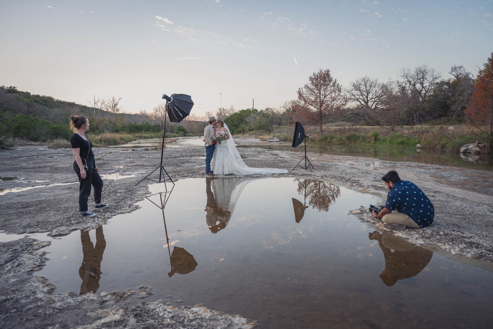 People are taking photos by a river with a bride and groom, one person is sitting on the ground, and equipment is set up for a photoshoot.