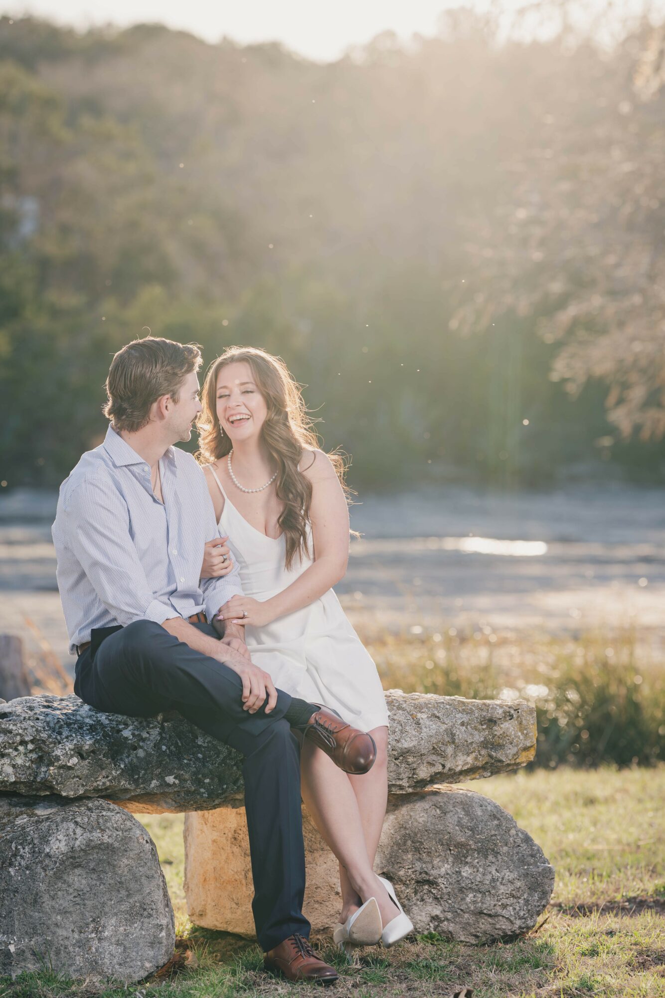 A couple sitting on a stone bench outdoors, smiling at each other with trees and sunlight in the background.
