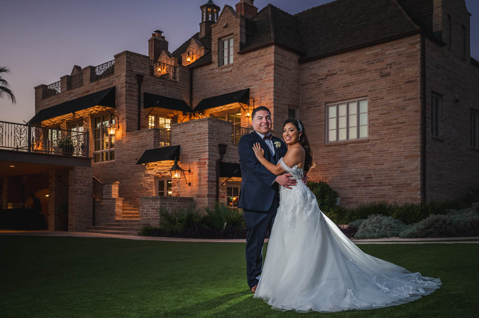 A couple in wedding attire standing on a lawn in front of a large, illuminated castle-like building at dusk.