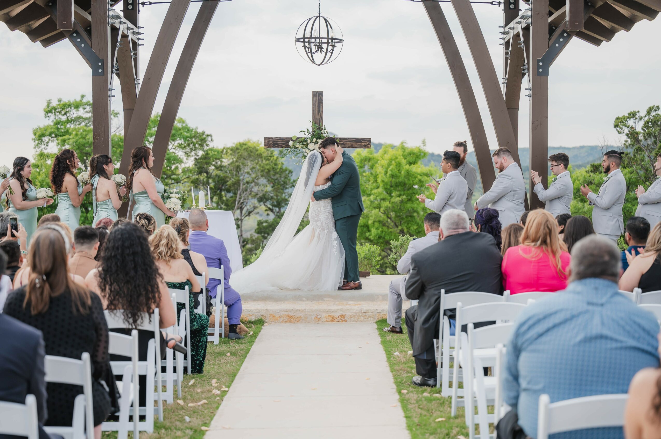 Couple kissing at outdoor wedding ceremony under wooden pavilion, guests seated on either side, greenery in background.