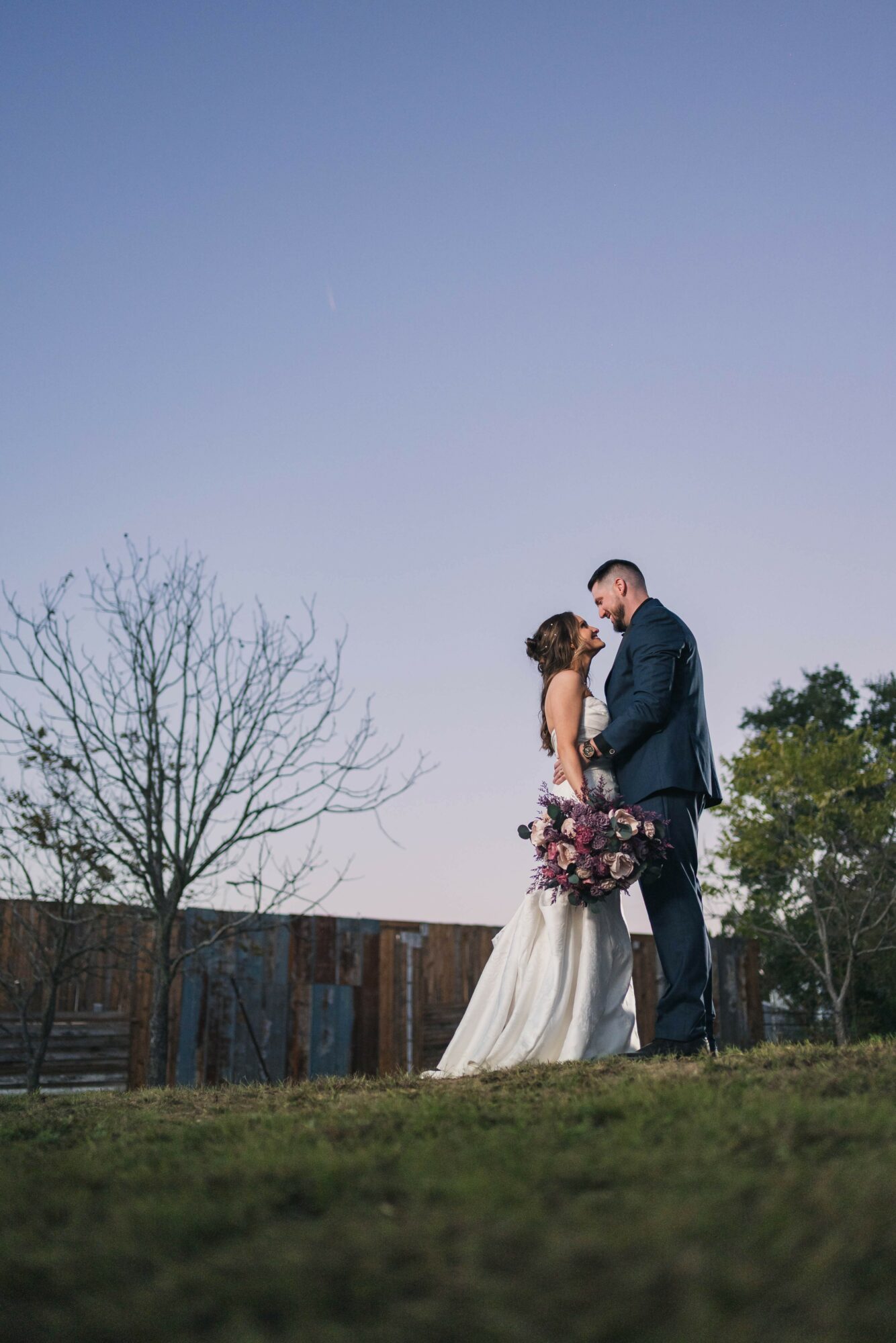 A bride and groom stand outdoors, holding a bouquet, with a clear sky and trees in the background.