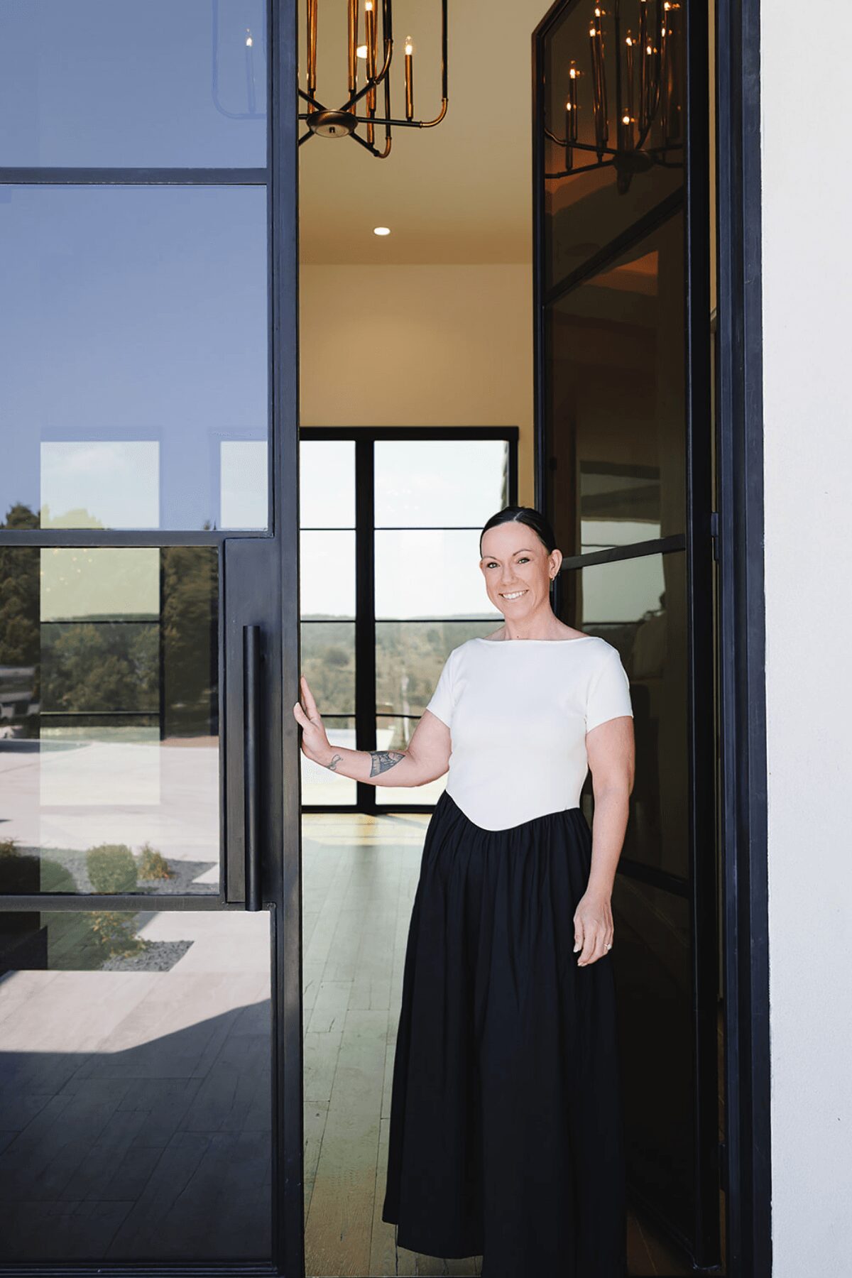 Woman standing in doorway, smiling, wearing a white top and black skirt, interior visible behind her.