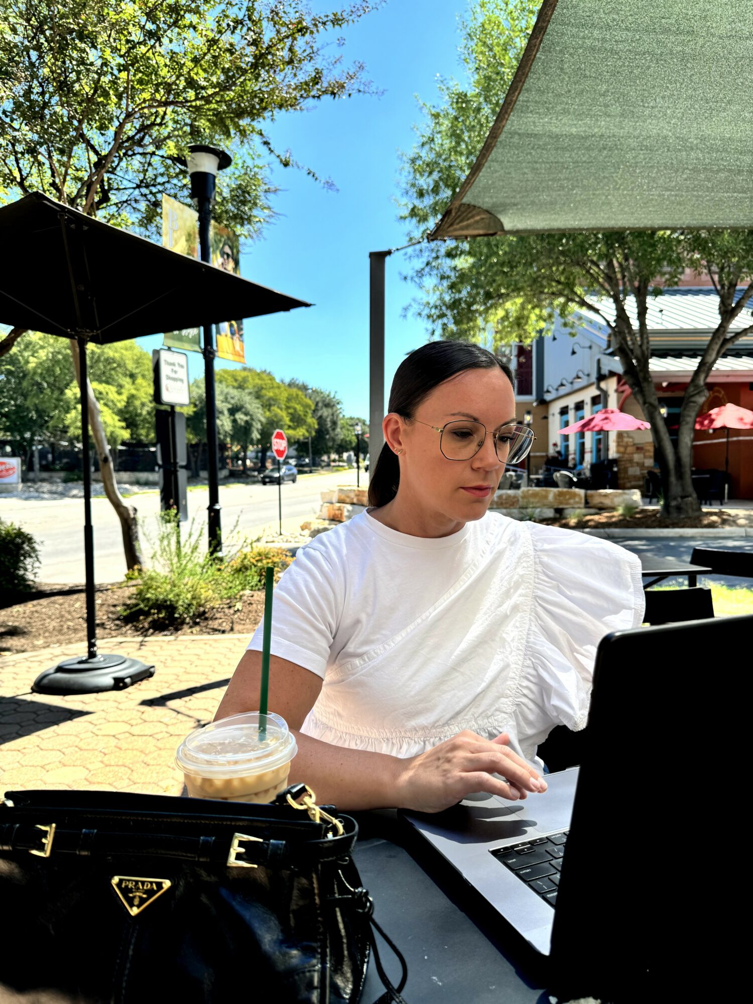 Woman sitting outdoors at a table with a laptop, iced coffee, and a black purse, under umbrellas on a sunny day.