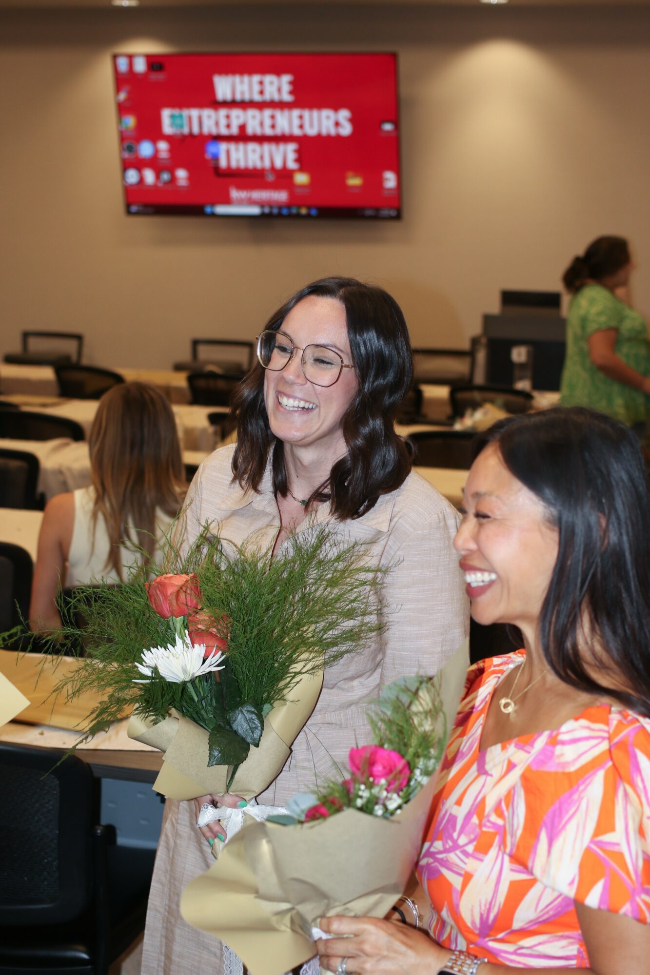 Two women smiling, holding flowers, in a conference room with a screen displaying 'WHERE ENTREPRENEURS THRIVE'.