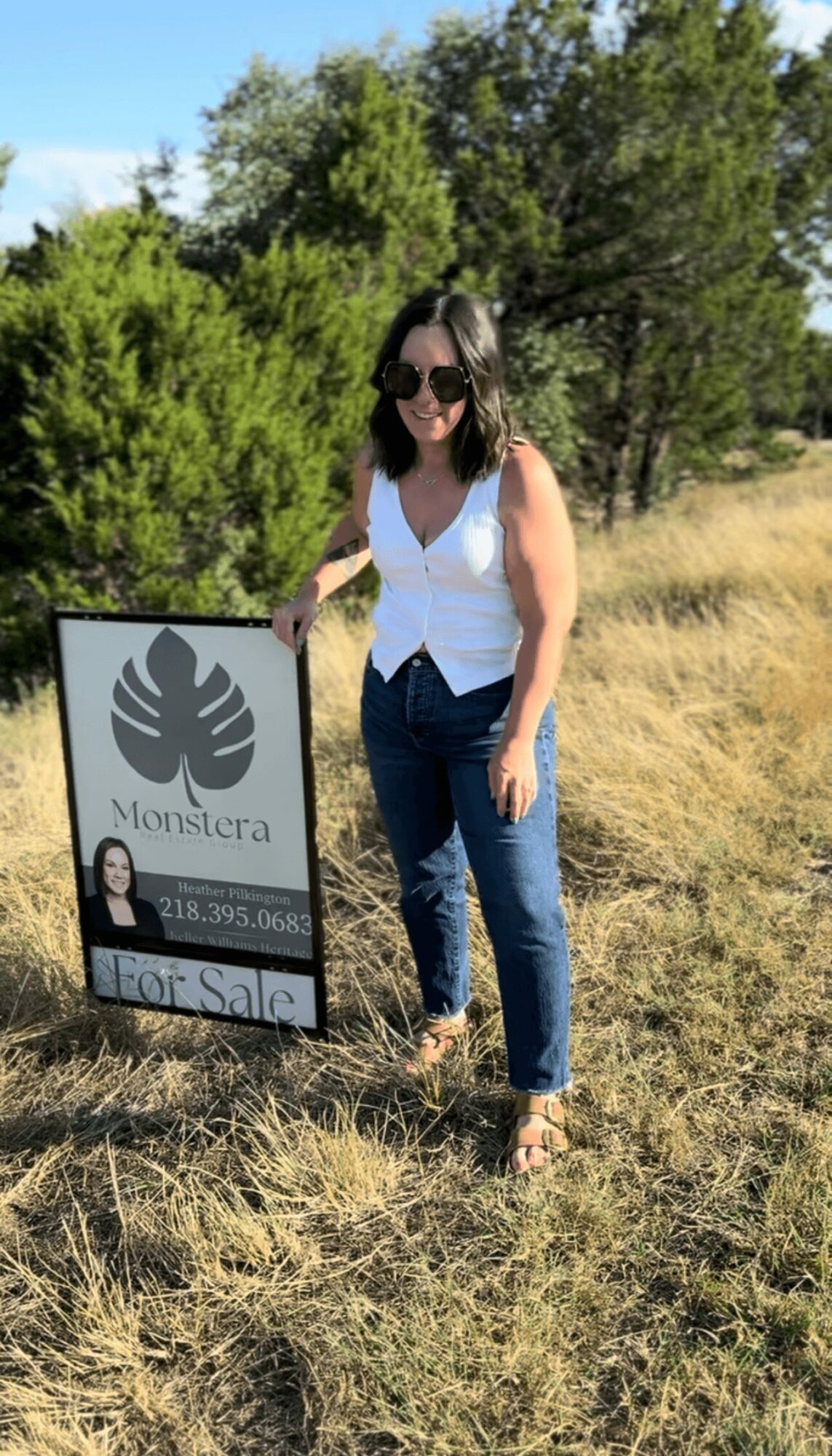 Woman standing outdoors next to a real estate sign, wearing sunglasses, white top, and jeans, with trees and grass in background.