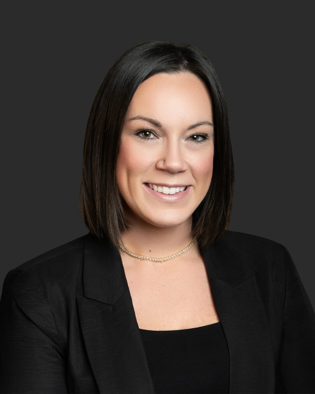 Portrait of a woman with shoulder-length dark hair, smiling, wearing a black blazer and top, against a dark background.