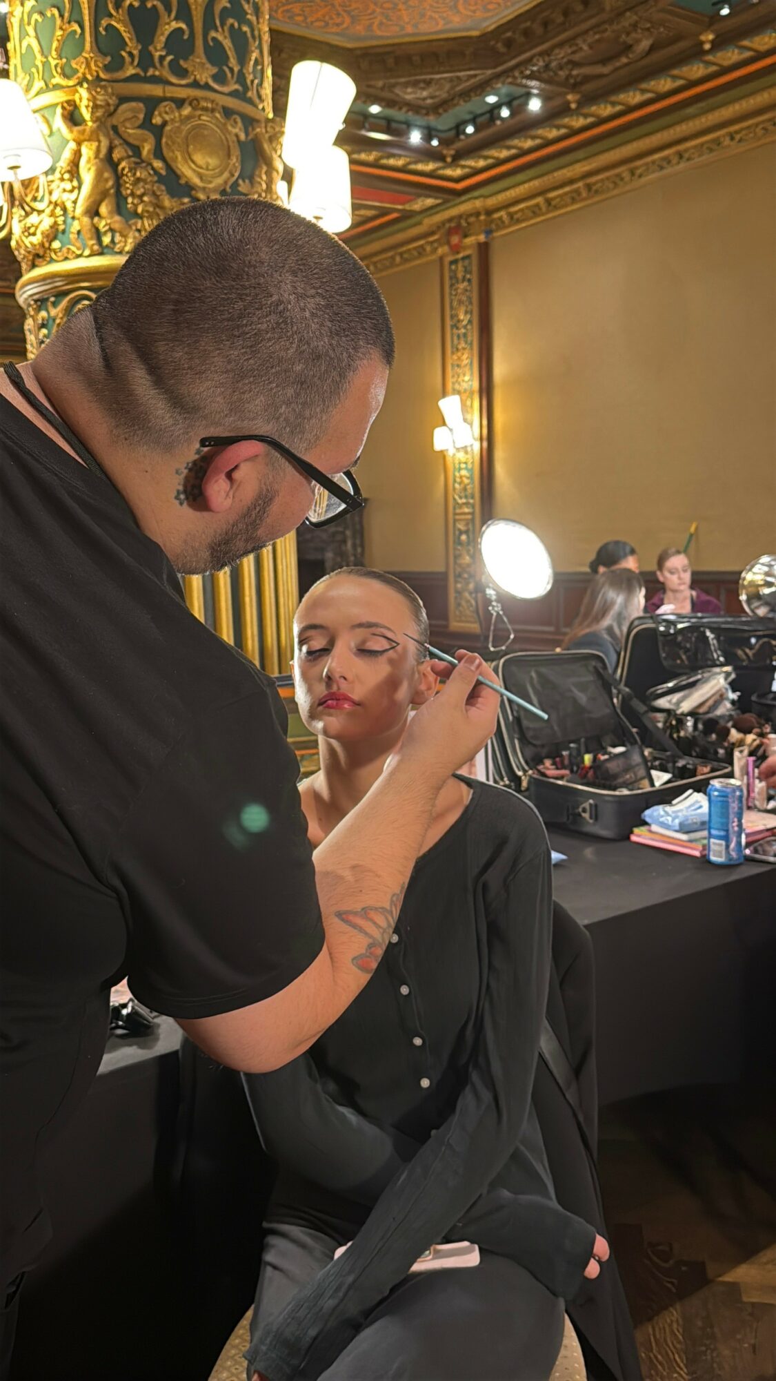 Makeup artist applying makeup to a woman in a black outfit, in a decorated room with ornate gold details and bright lights.