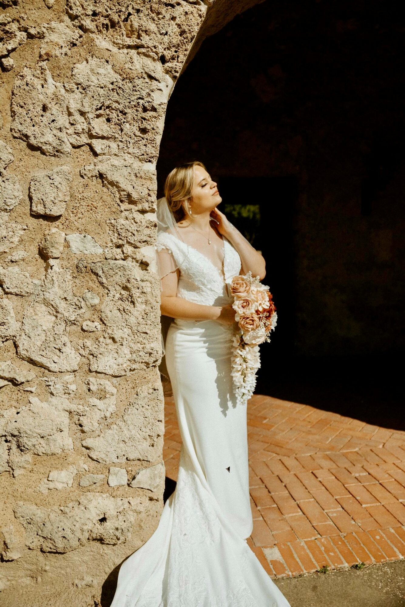 Woman in wedding dress holding a bouquet, standing against a stone wall with an archway, sunlight on her face.