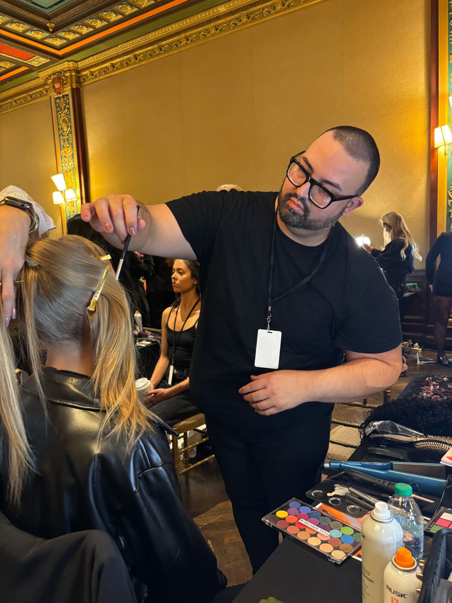 Man with glasses and a beard applying makeup to a young girl with blonde hair in a black leather jacket, in a decorated room.