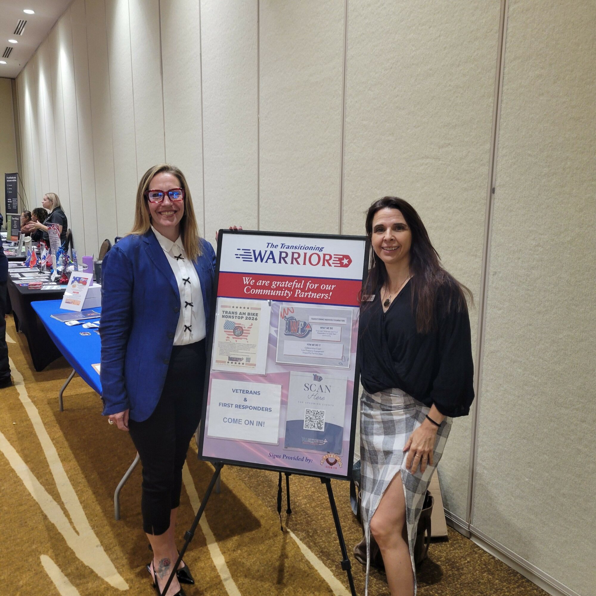 Two women standing next to a display board at an indoor event, with a table of items in front of them.