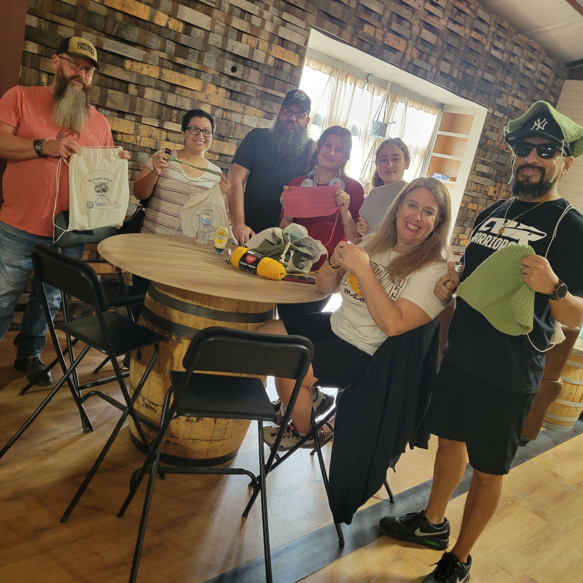 Group of six people standing around a table with a cake, smiling indoors with brick walls and wooden flooring.