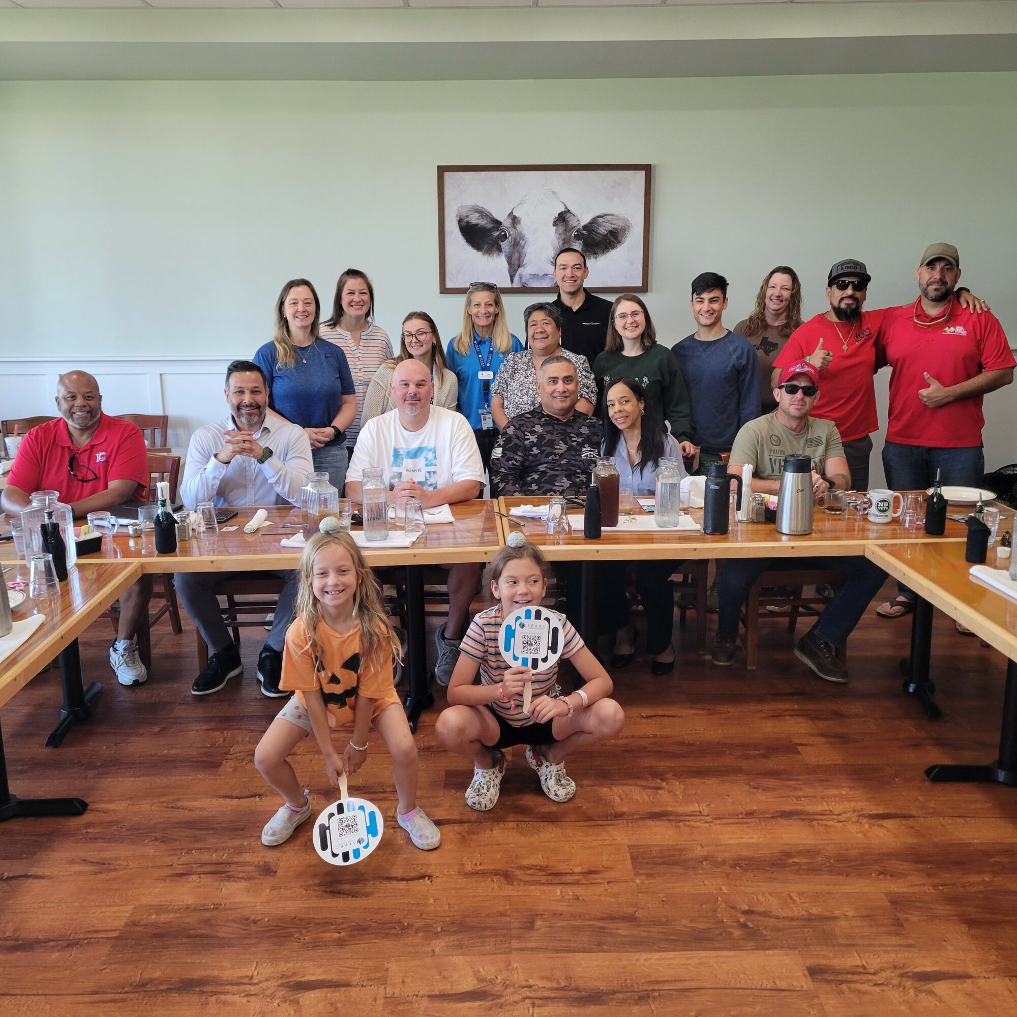 Group of 16 people in a room with a wooden floor, sitting and standing around a table, some holding devices, with a framed picture of butterflies on the wall.