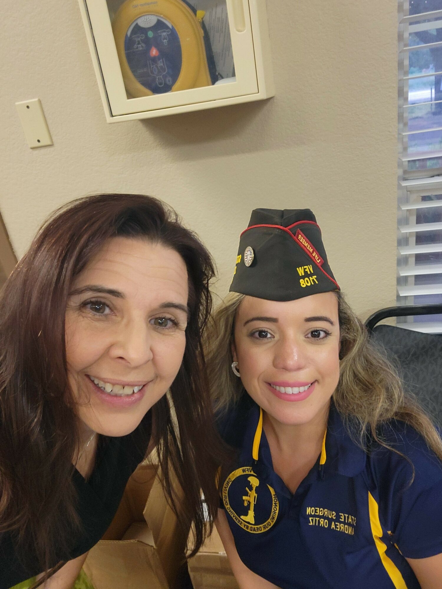 Two women smiling, one wearing a navy uniform and cap, the other with long brown hair, in an indoor setting.