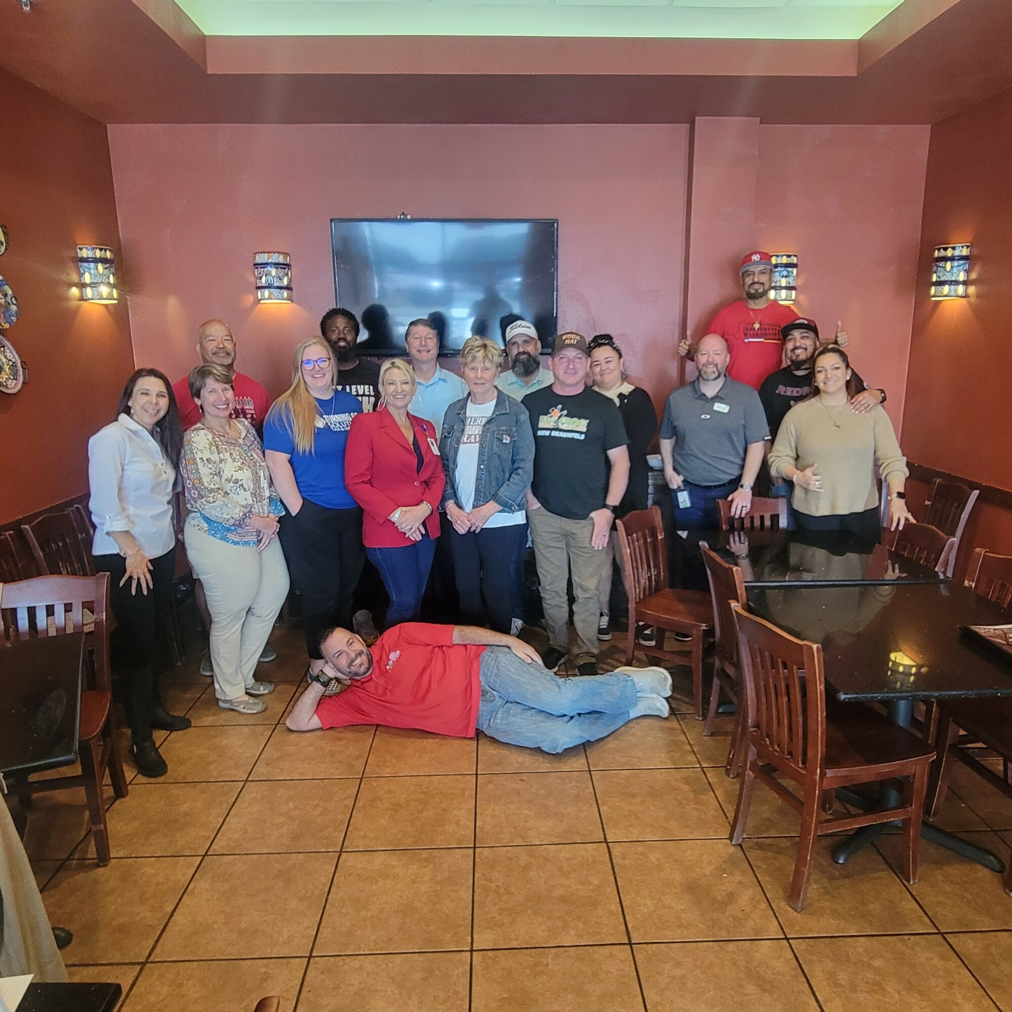 Group of people standing in a restaurant with one person lying on the floor in front of them, all smiling.