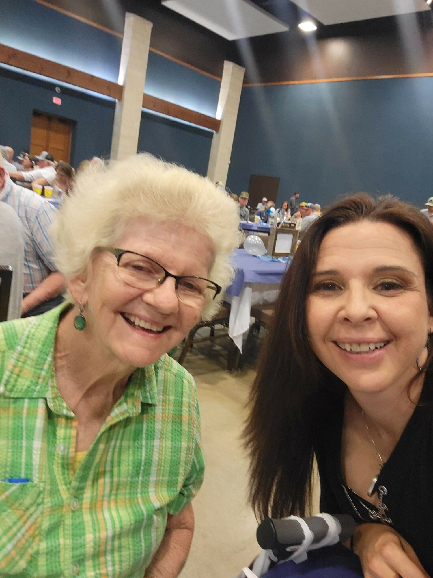Two women smiling, one with glasses and white hair, the other with long dark hair, in a room with tables and chairs.