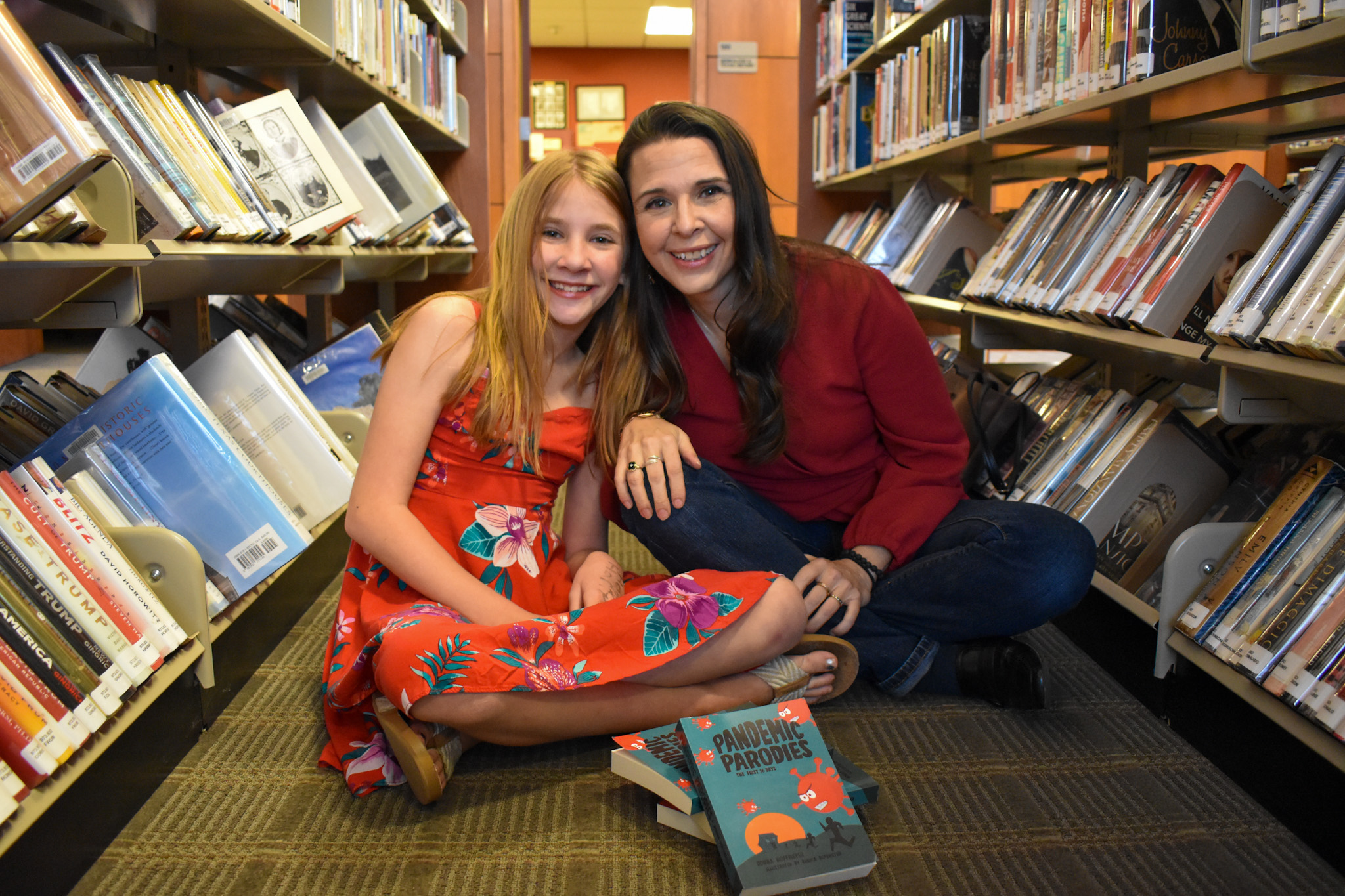 Two people sitting on the floor in a library aisle, smiling, with books around them and a book on the floor.