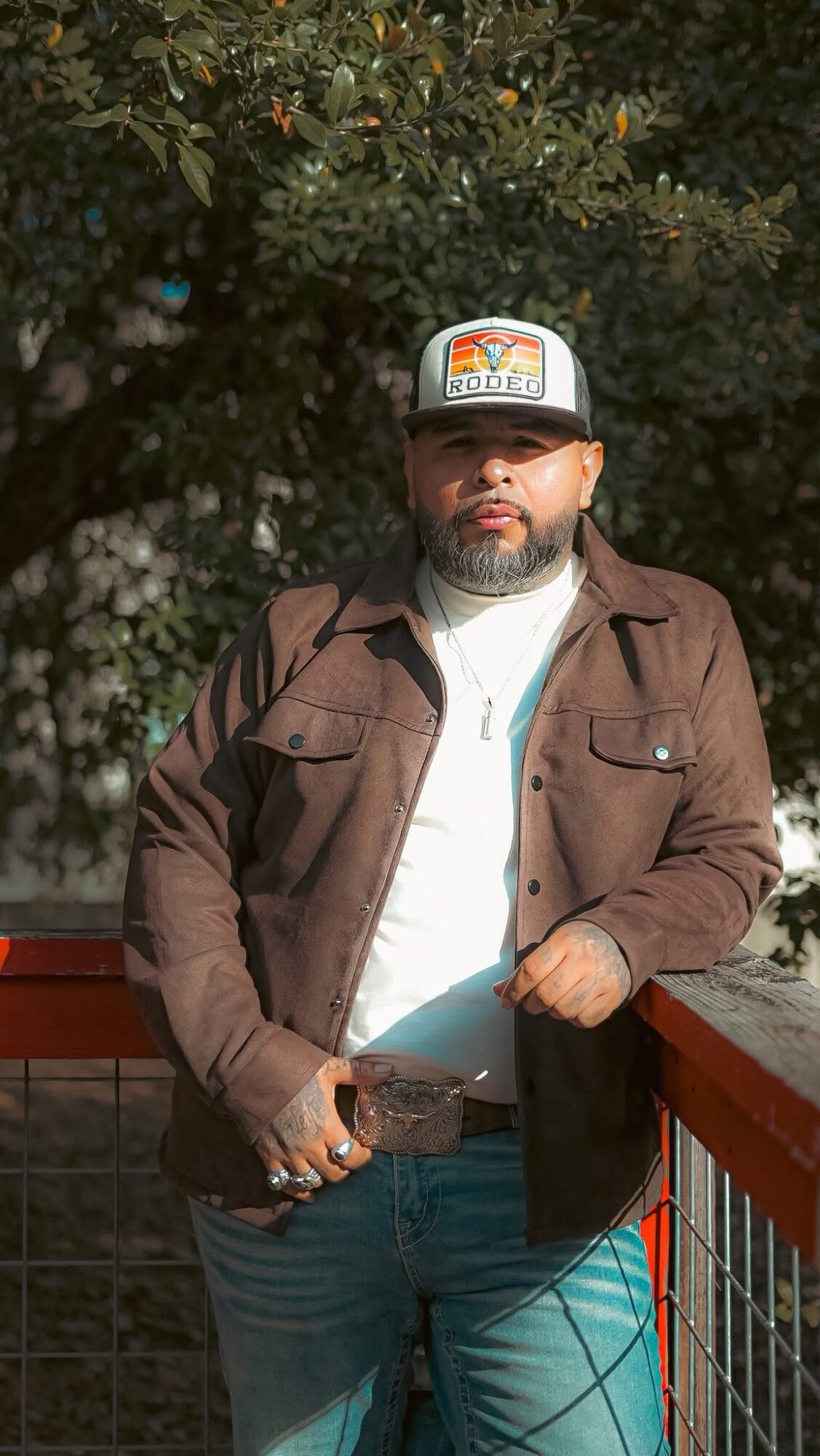 Man with beard wearing a cap, brown jacket, and white shirt, standing outdoors near a railing with trees in background.