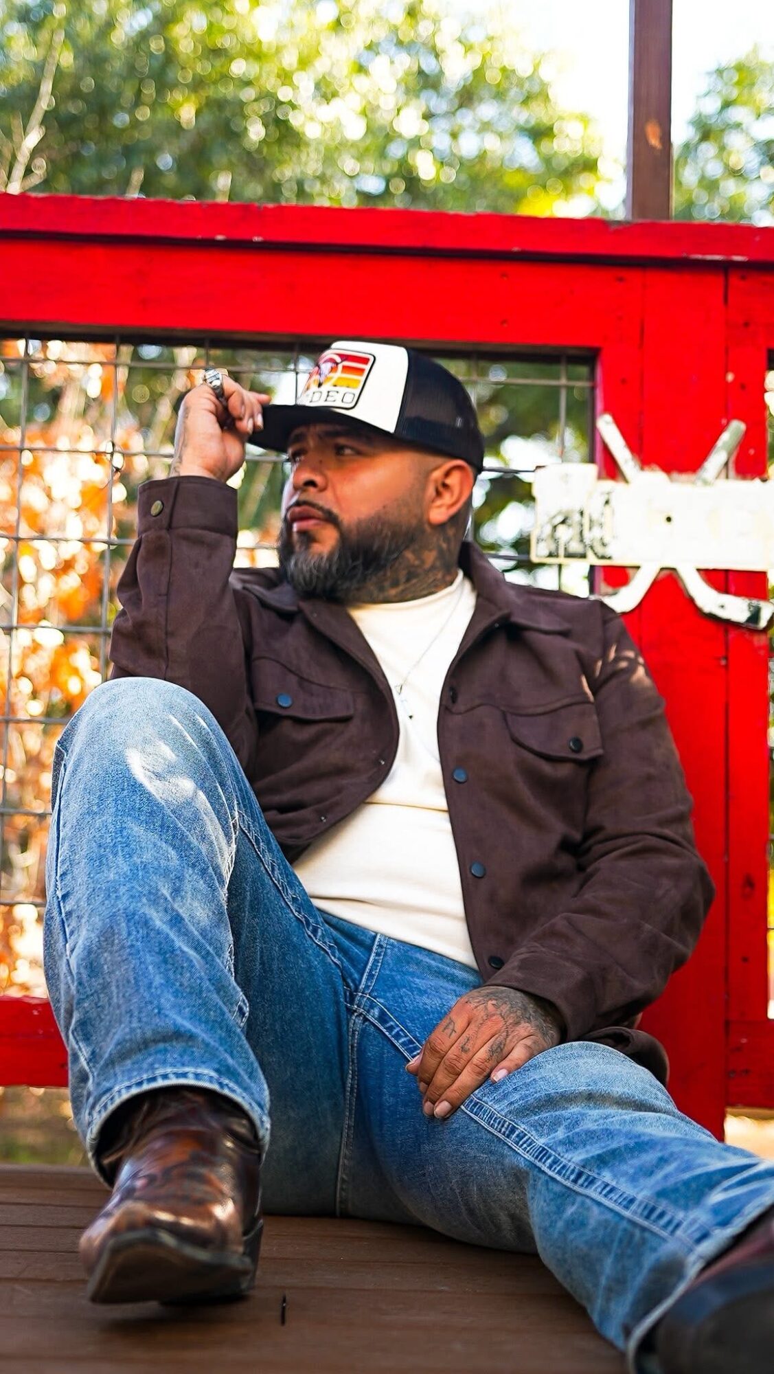 Man sitting outdoors in front of a red structure, wearing a black jacket, white shirt, jeans, and a cap, looking to the side.
