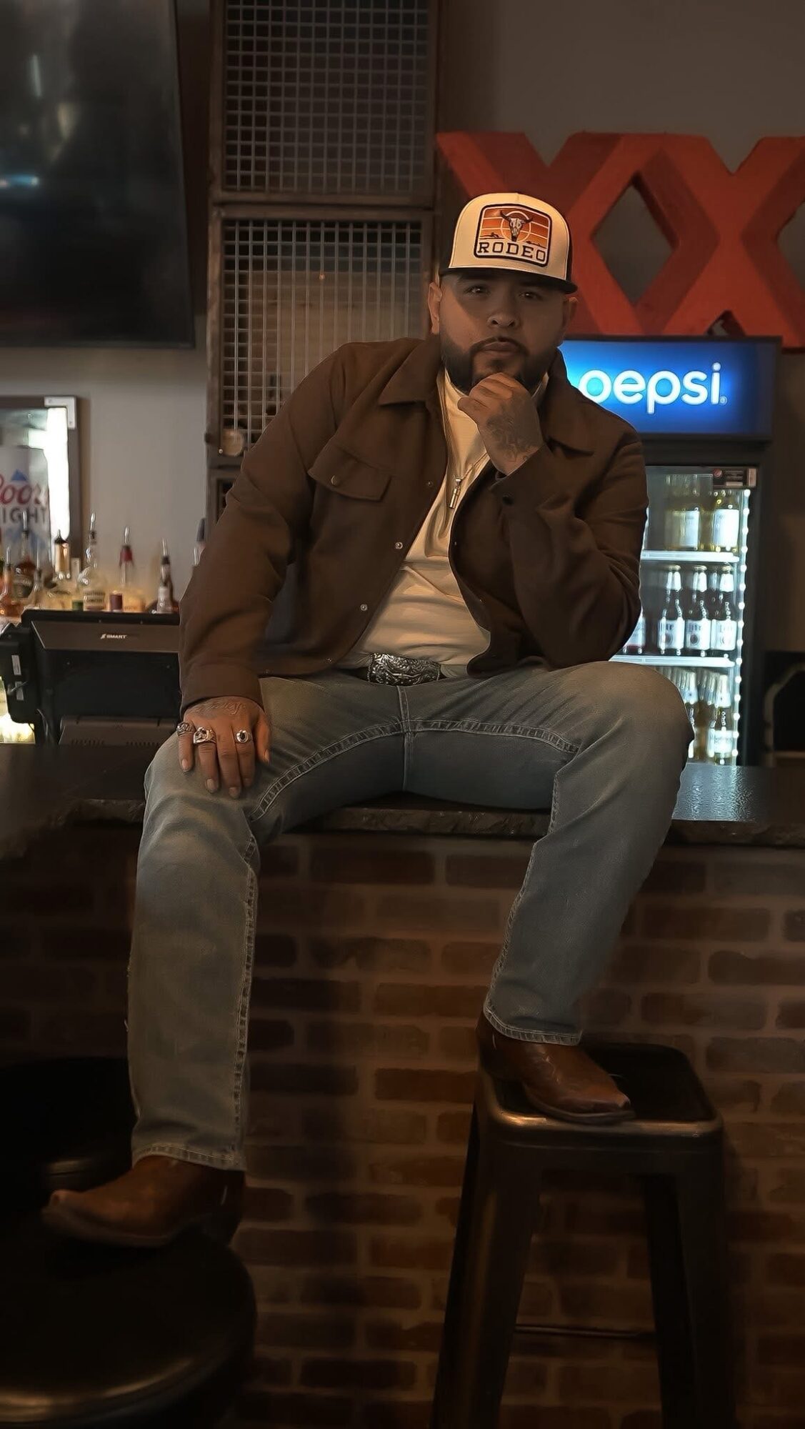 Man sitting on a bar counter with a Pepsi sign behind him, wearing a cap, jacket, and jeans, resting his chin on his hand.