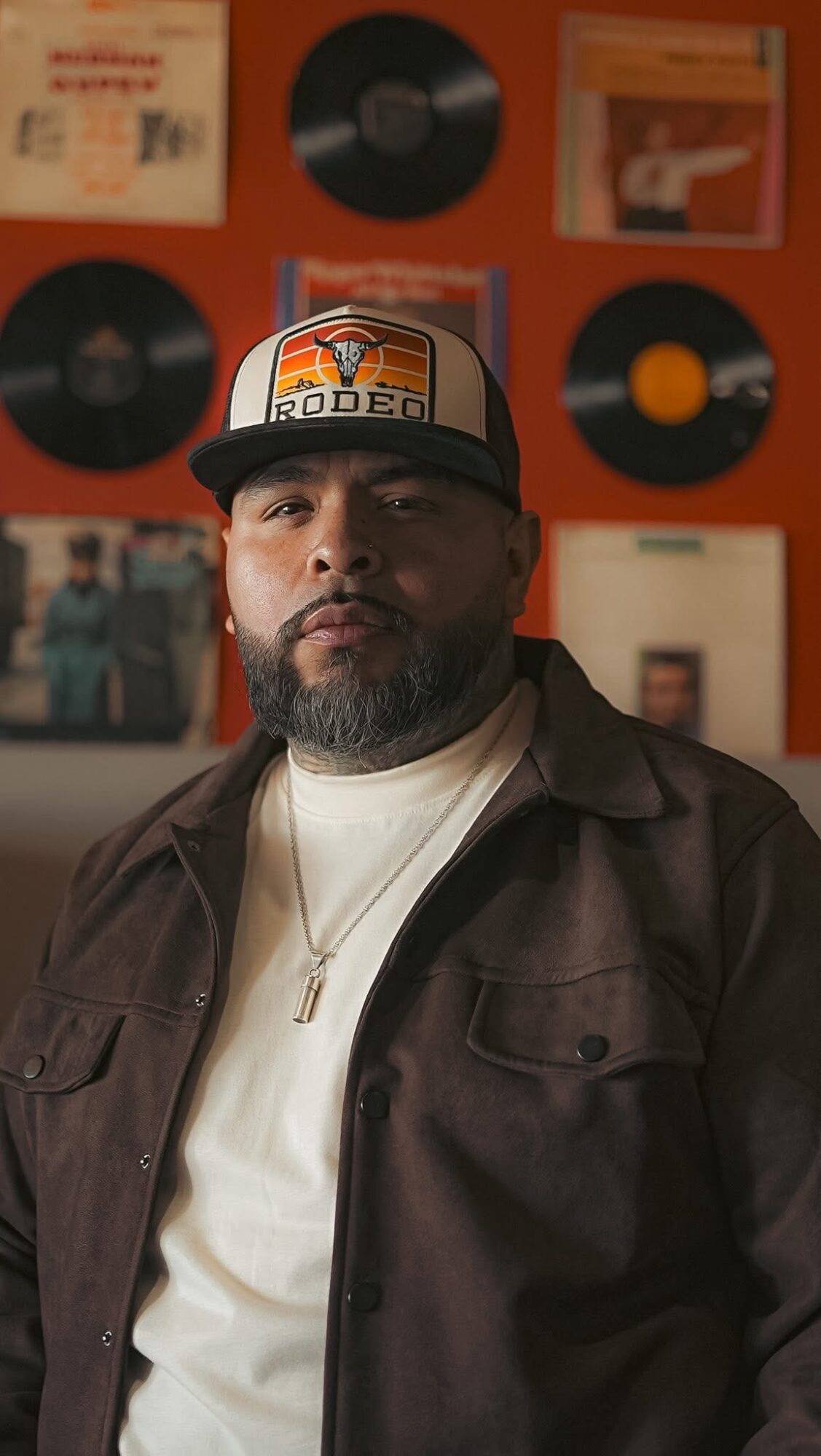 Man with beard wearing a cap and dark jacket, standing in front of a wall with vinyl records and photos.