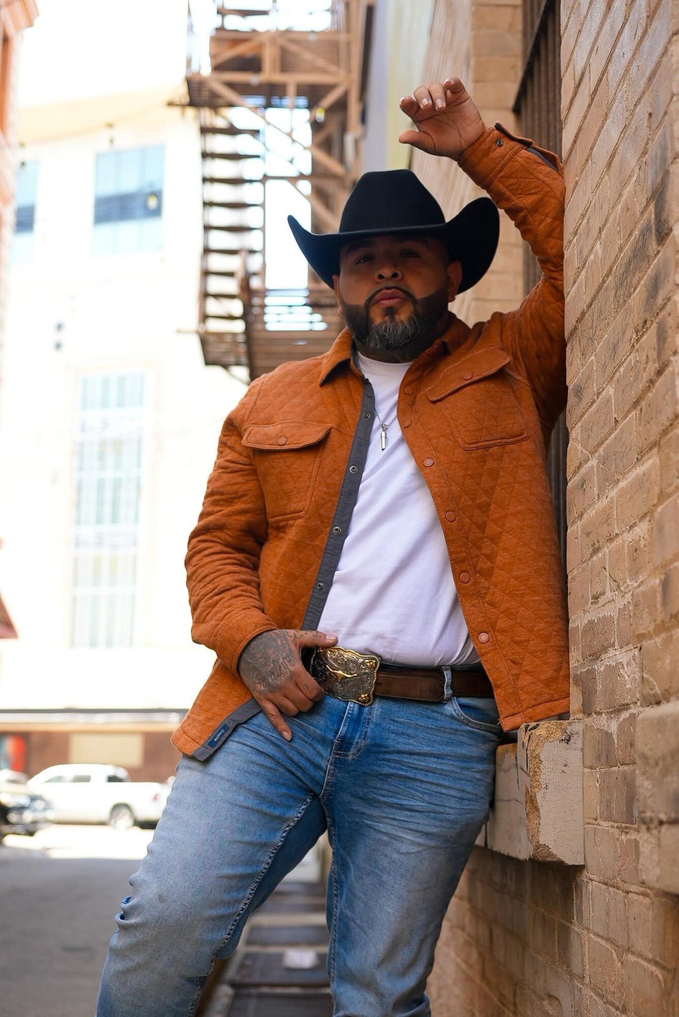 Man in cowboy hat and orange jacket leaning against brick wall outdoors, with city buildings in background.