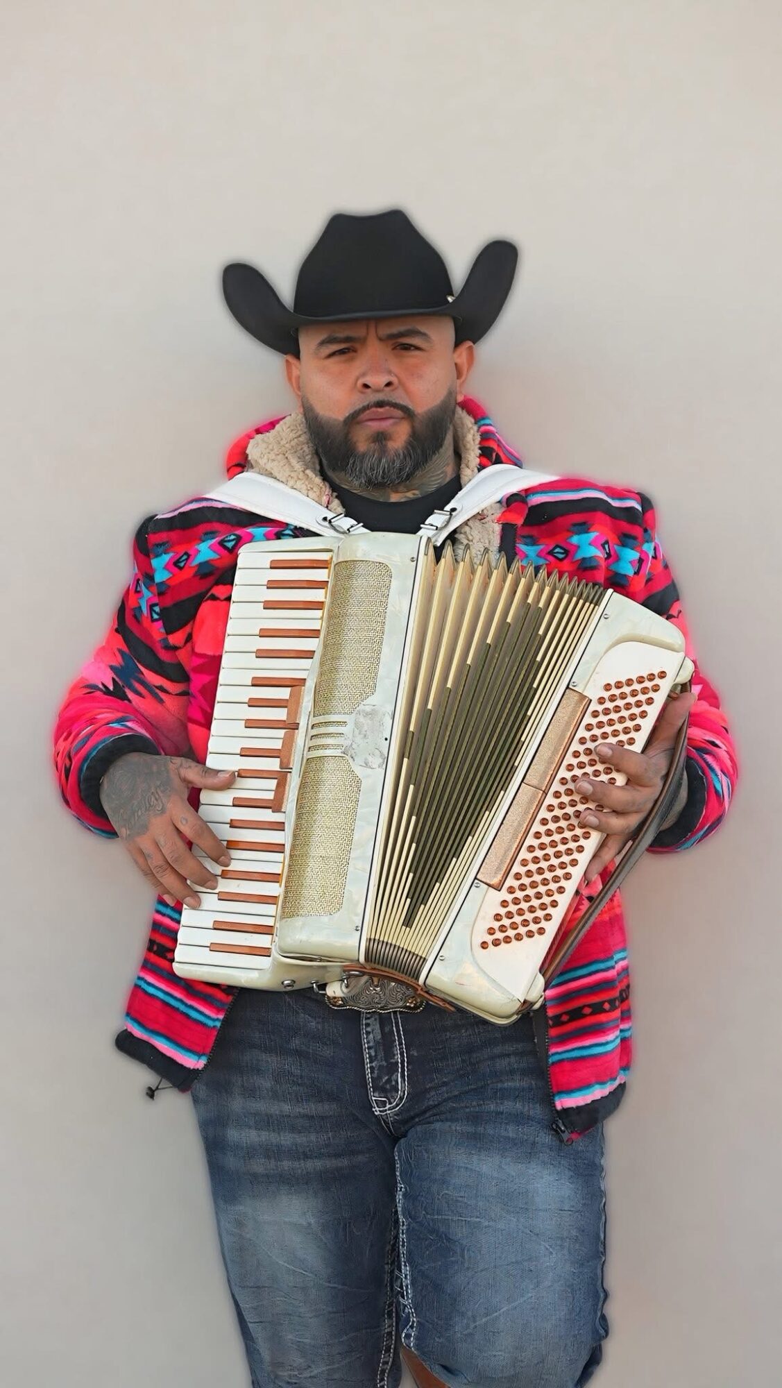 Man wearing a cowboy hat and colorful jacket holding an accordion, standing against a plain wall.