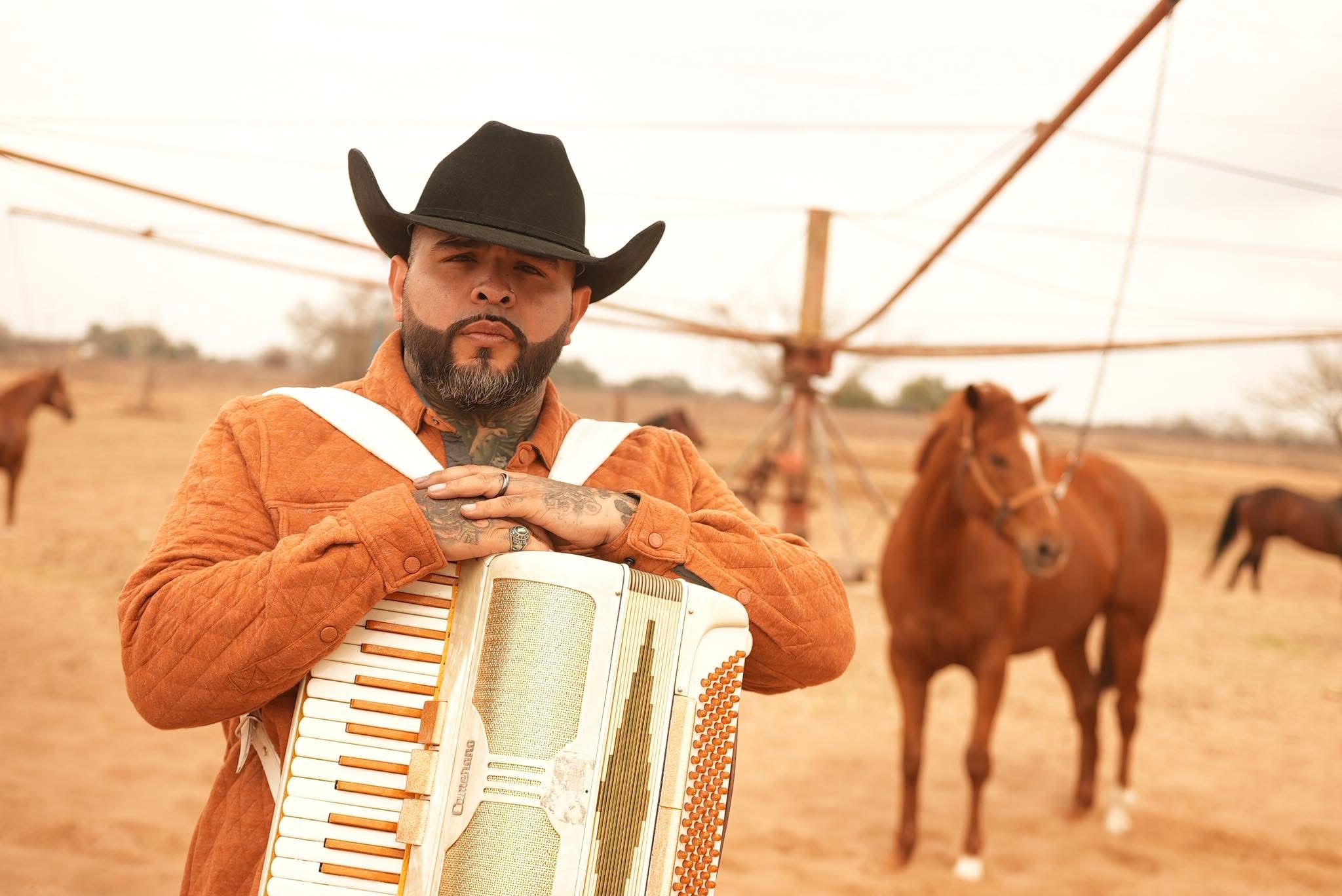 Man wearing cowboy hat and orange jacket holding an accordion outdoors with horses in the background.