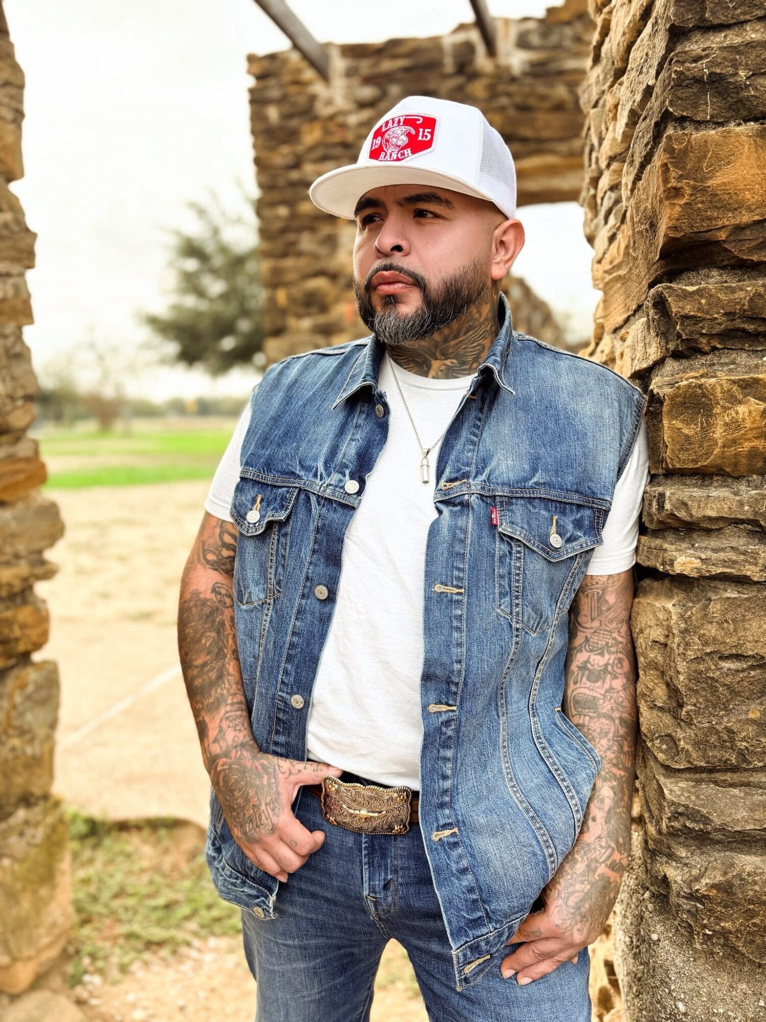 Man with a beard and tattoos wearing a white cap, denim vest, and white t-shirt, standing between stone walls outdoors.
