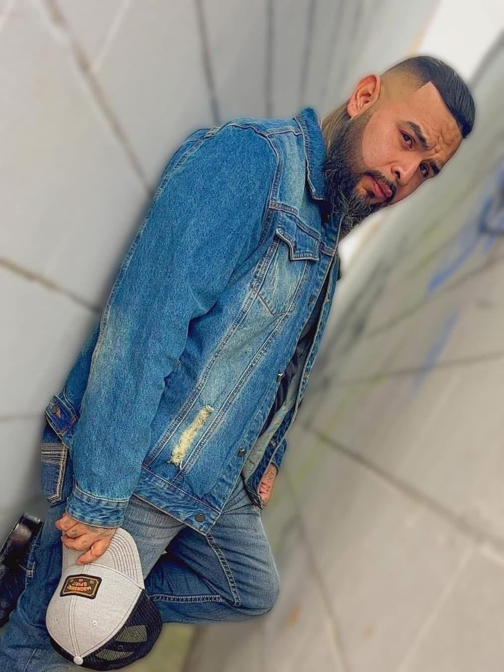 Man leaning against a tiled wall, wearing a denim jacket and holding a white cap, looking at the camera.